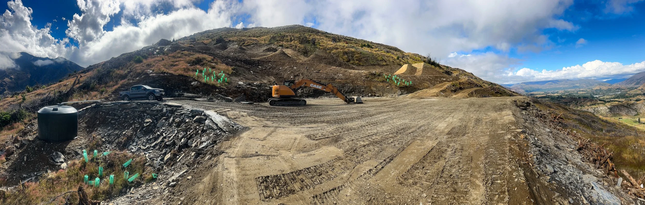 Construction site on a hillside with a black truck, an orange CASE excavator, green plant protectors, and wide view of mountains and partly cloudy sky.