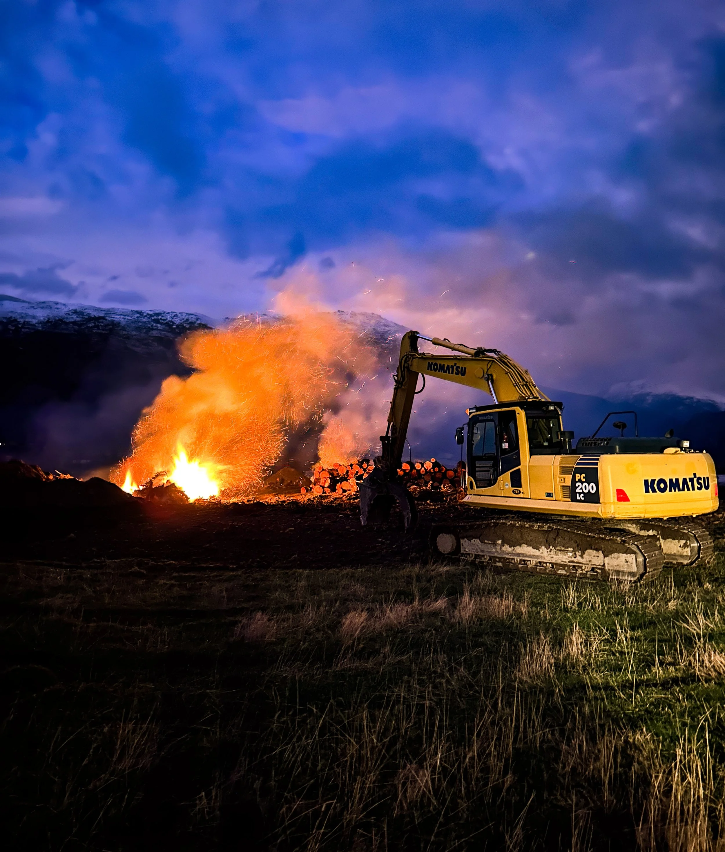 A yellow Komatsu excavator working on a pile of logs near a fire in a mountainous landscape during dusk or dawn.