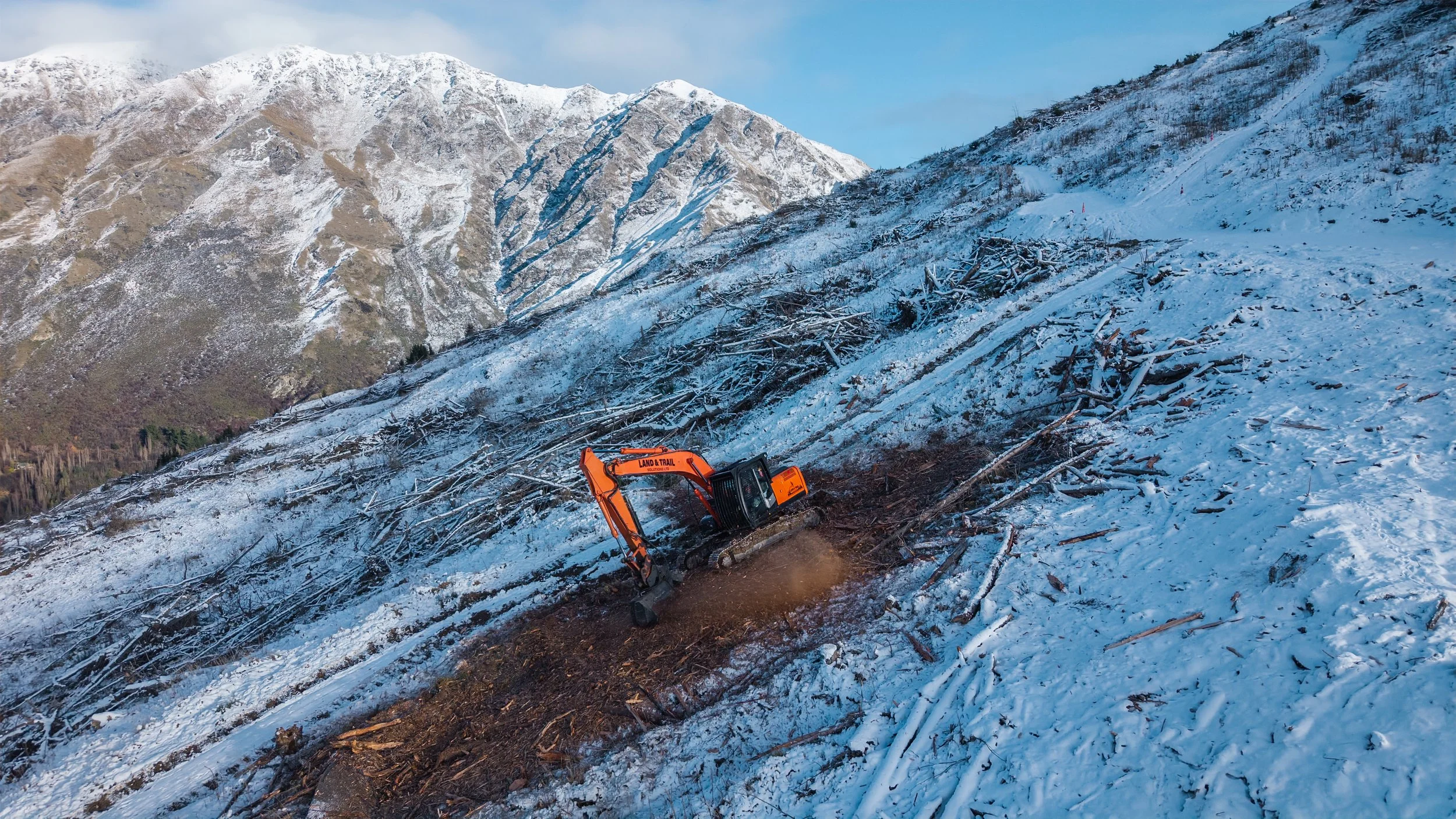An orange excavator clearing fallen trees from a snow-covered mountainside, with snow-covered mountains in the background.