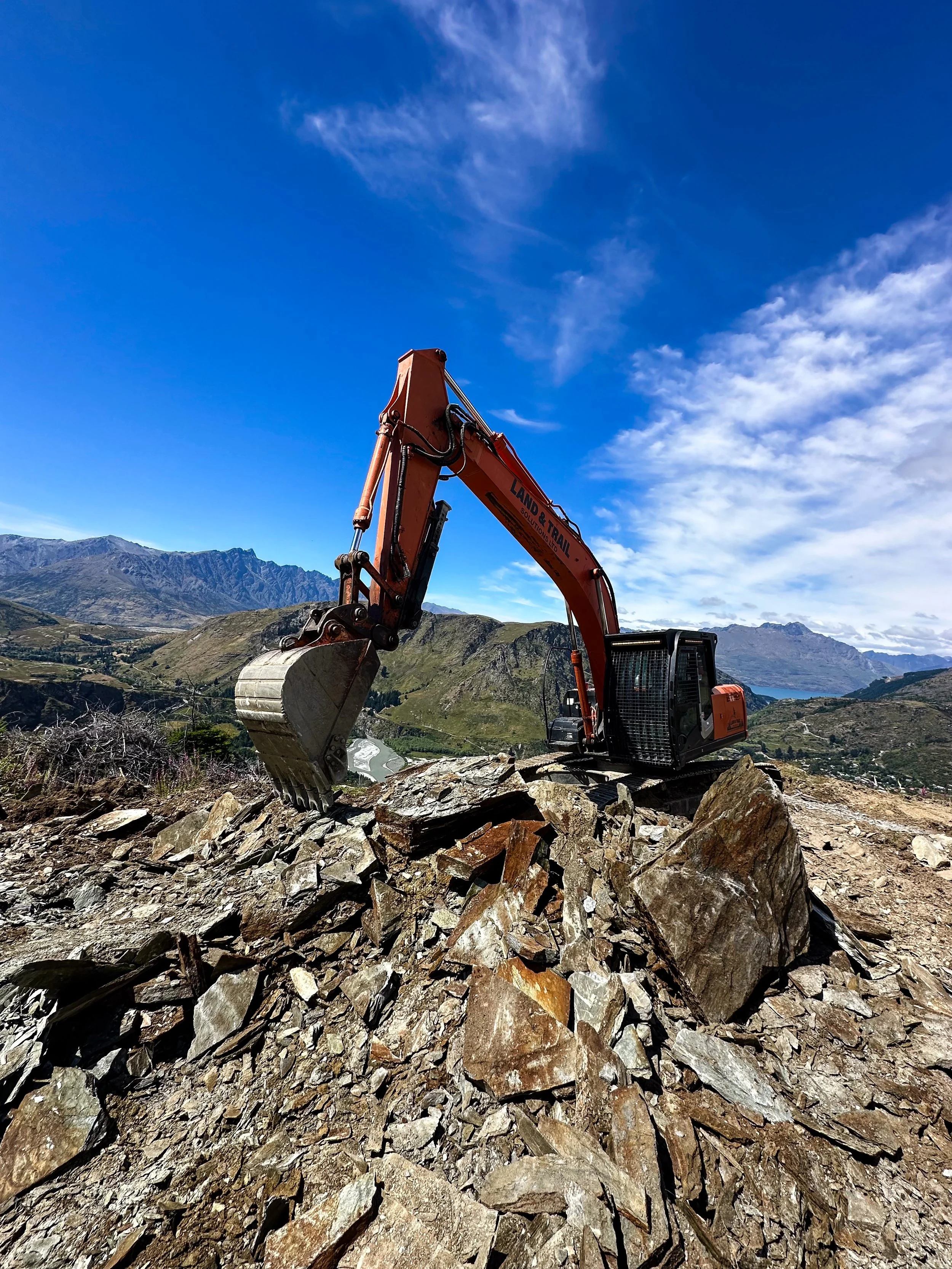 An orange excavator working on a rocky hillside with mountains and a blue sky in the background.