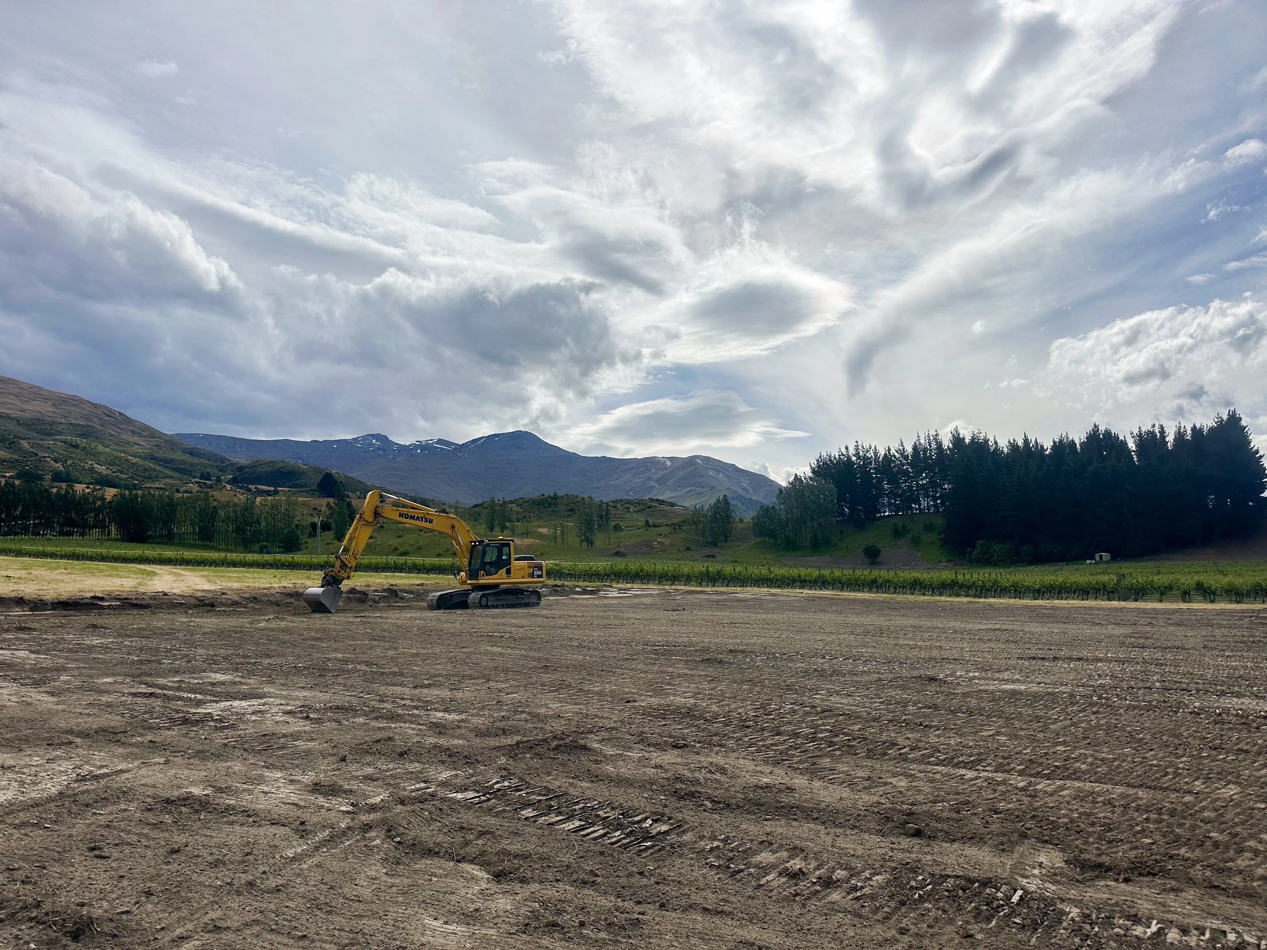 A yellow construction excavator working on a large open field with mountains and cloudy sky in the background.