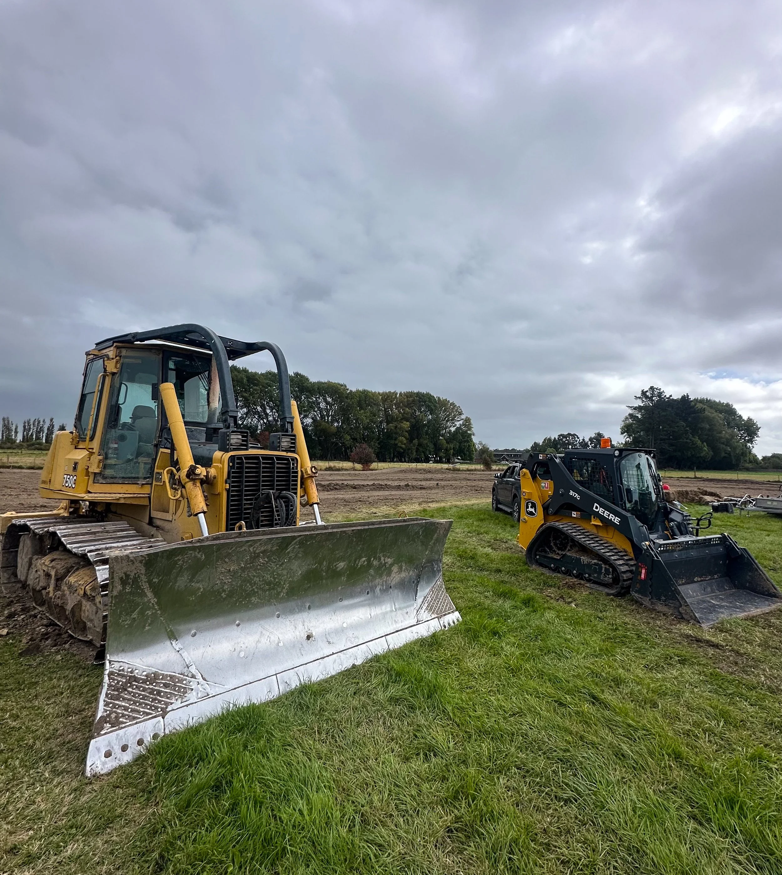 Construction site with a yellow bulldozer and a black skid steer loader on grassy terrain under cloudy sky.