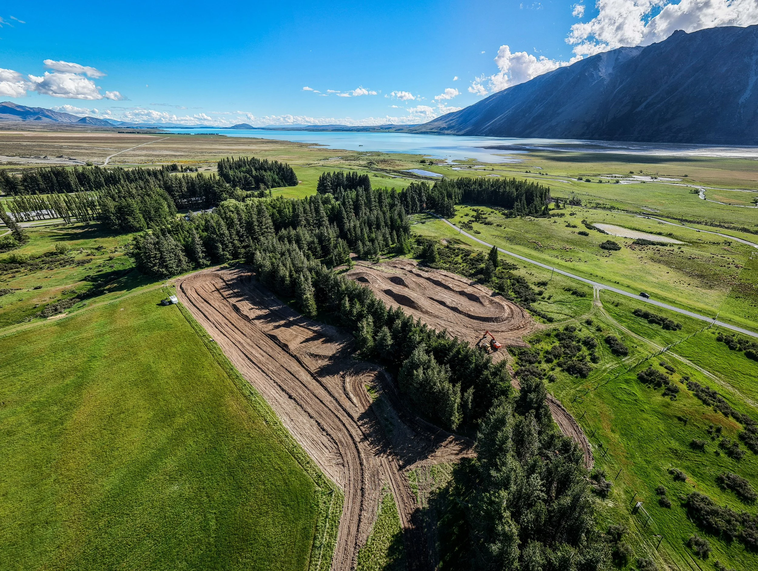 Aerial view of a landscape with a lake, mountains, green fields, and a construction site with cleared land and trees. Motocross track, outdoors mx track nz, new zealand motocross