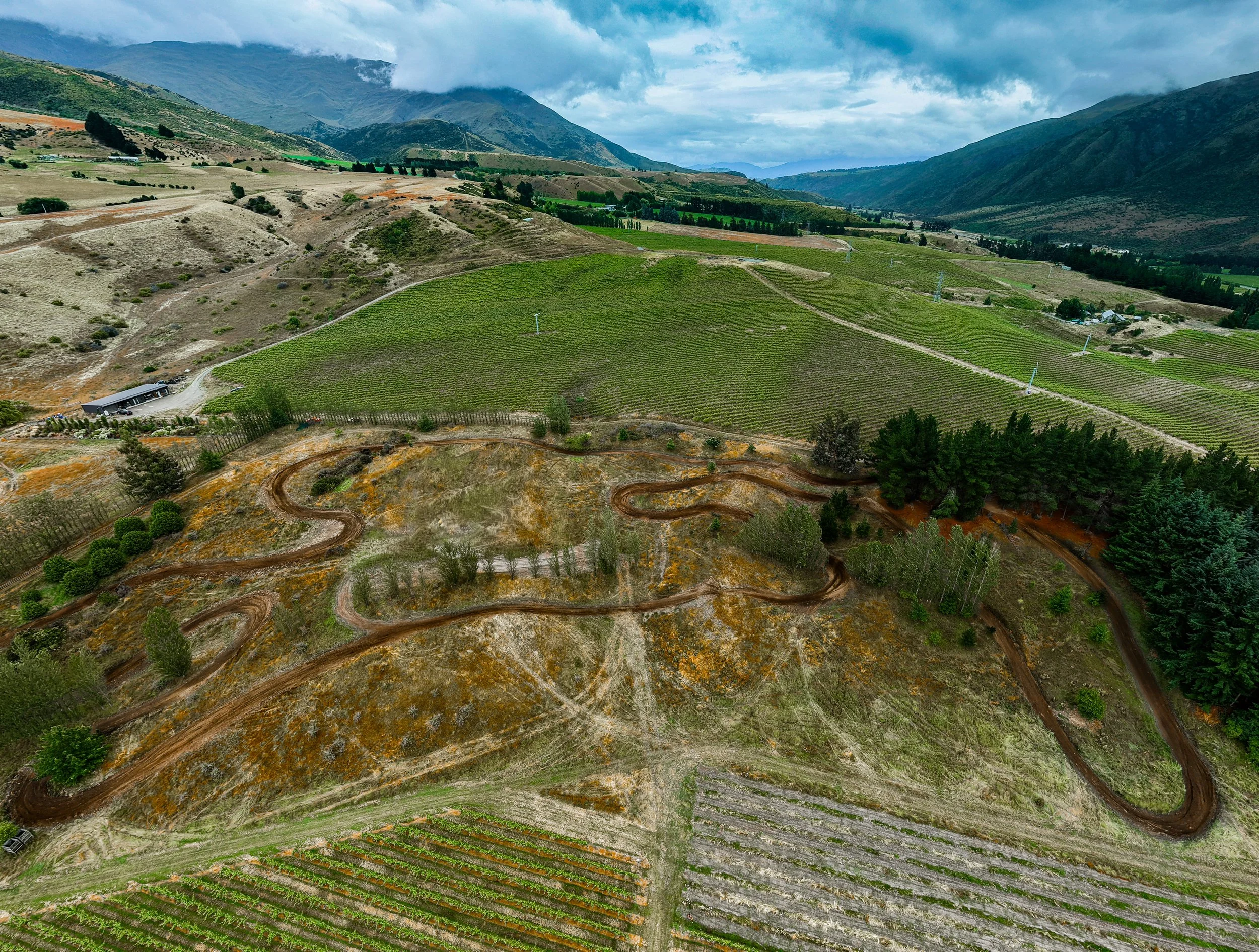 Aerial view of a landscape with winding dirt road through hills and vineyard fields in a valley surrounded by mountains and cloudy sky. Outdoor motocross track, mx park nz, motocross jumps, track building
