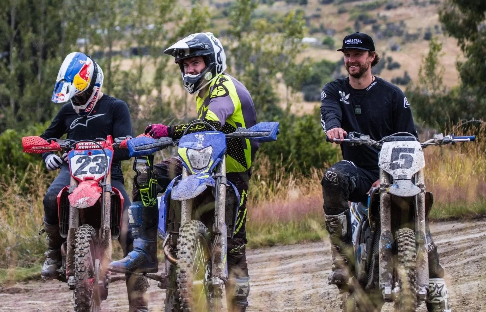 Three motocross riders on dirt bikes, riding on a dirt trail outdoors with trees and hills in the background, appears muddy and enjoying their ride.