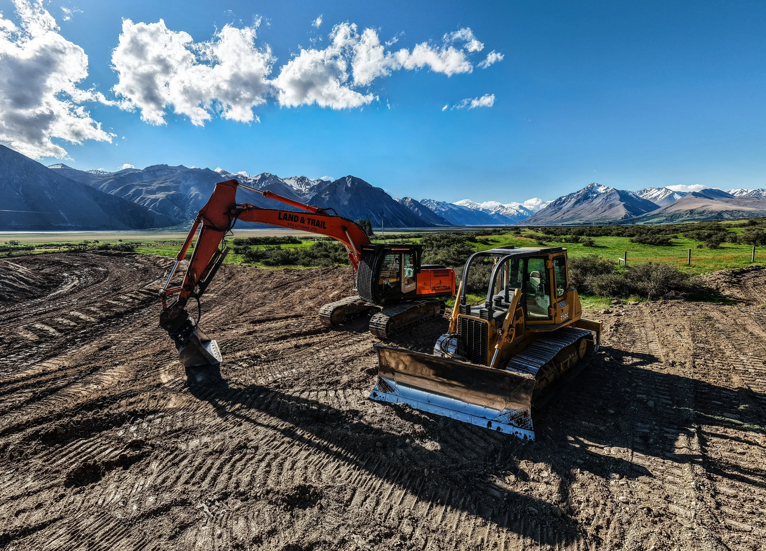 Construction site with an orange excavator and a yellow bulldozer working on the dirt. Snow-capped mountains and green fields in the background under a partly cloudy sky.