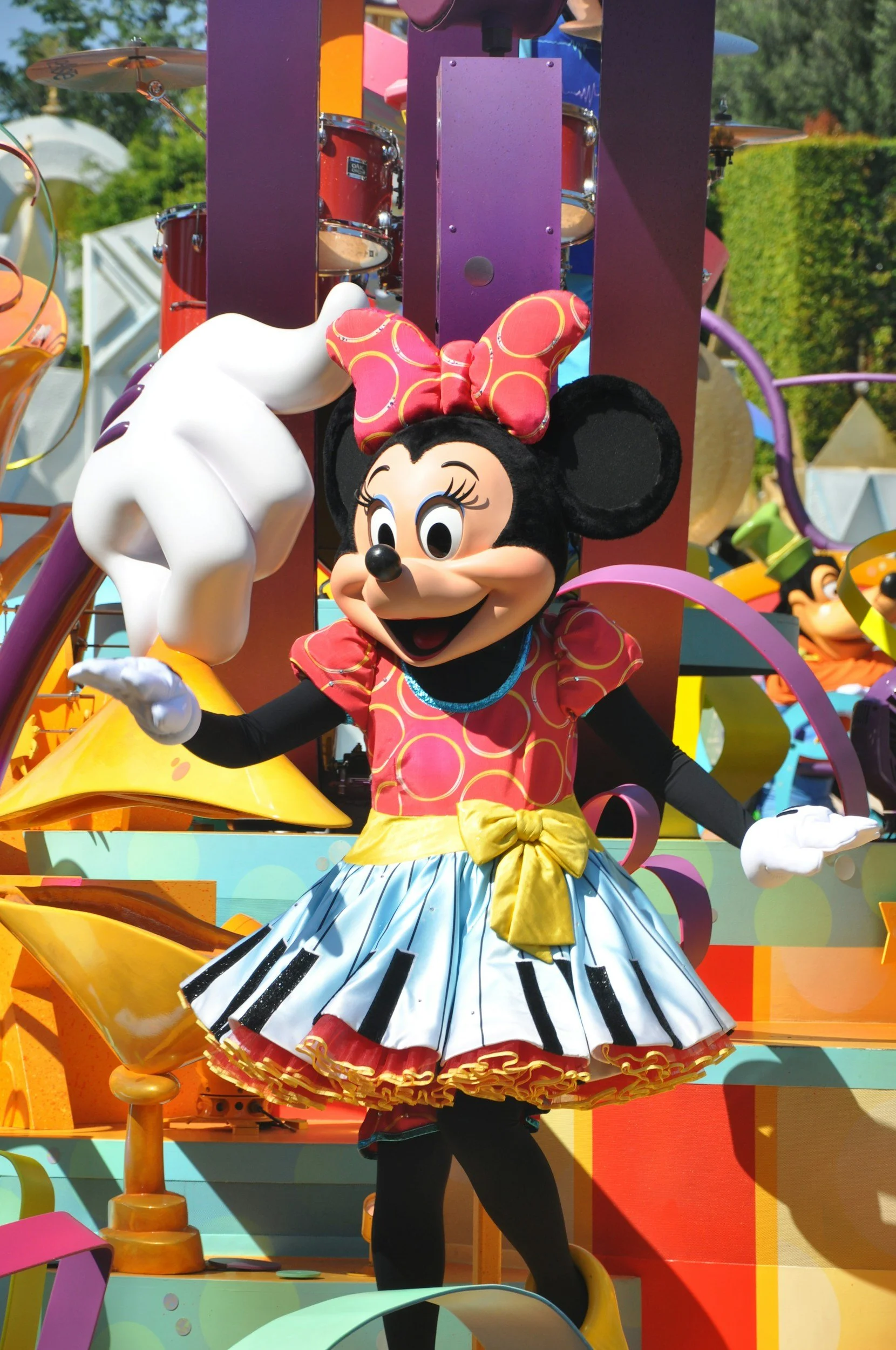 Costumed character of Minnie Mouse on a colorful parade float, wearing a red dress with yellow and black accents, a large red polka-dot bow, and black ears, with a whimsical and vibrant background featuring various amusement park decorations.