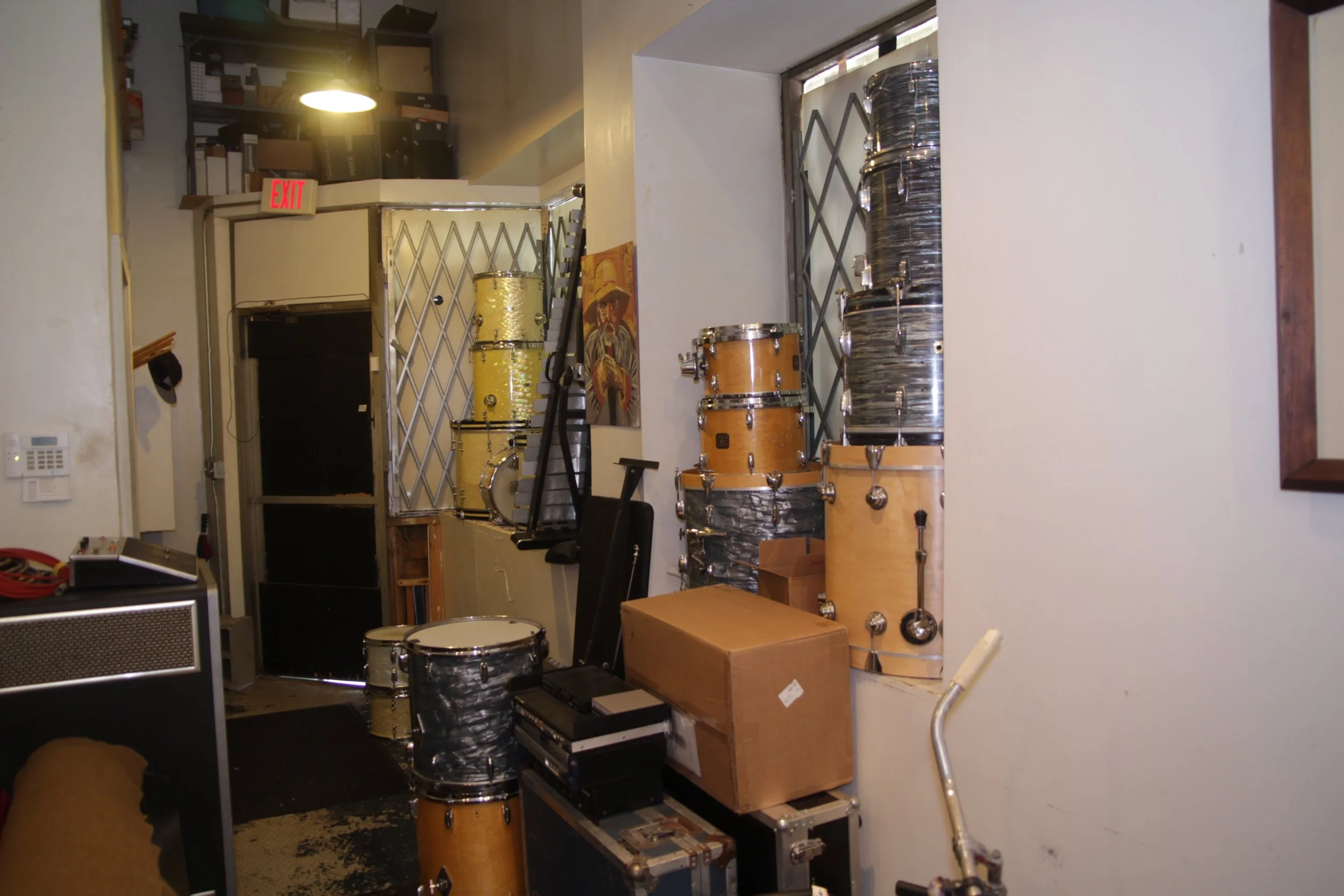 Room with stacked drums, cardboard boxes, and equipment cases near an exit sign.