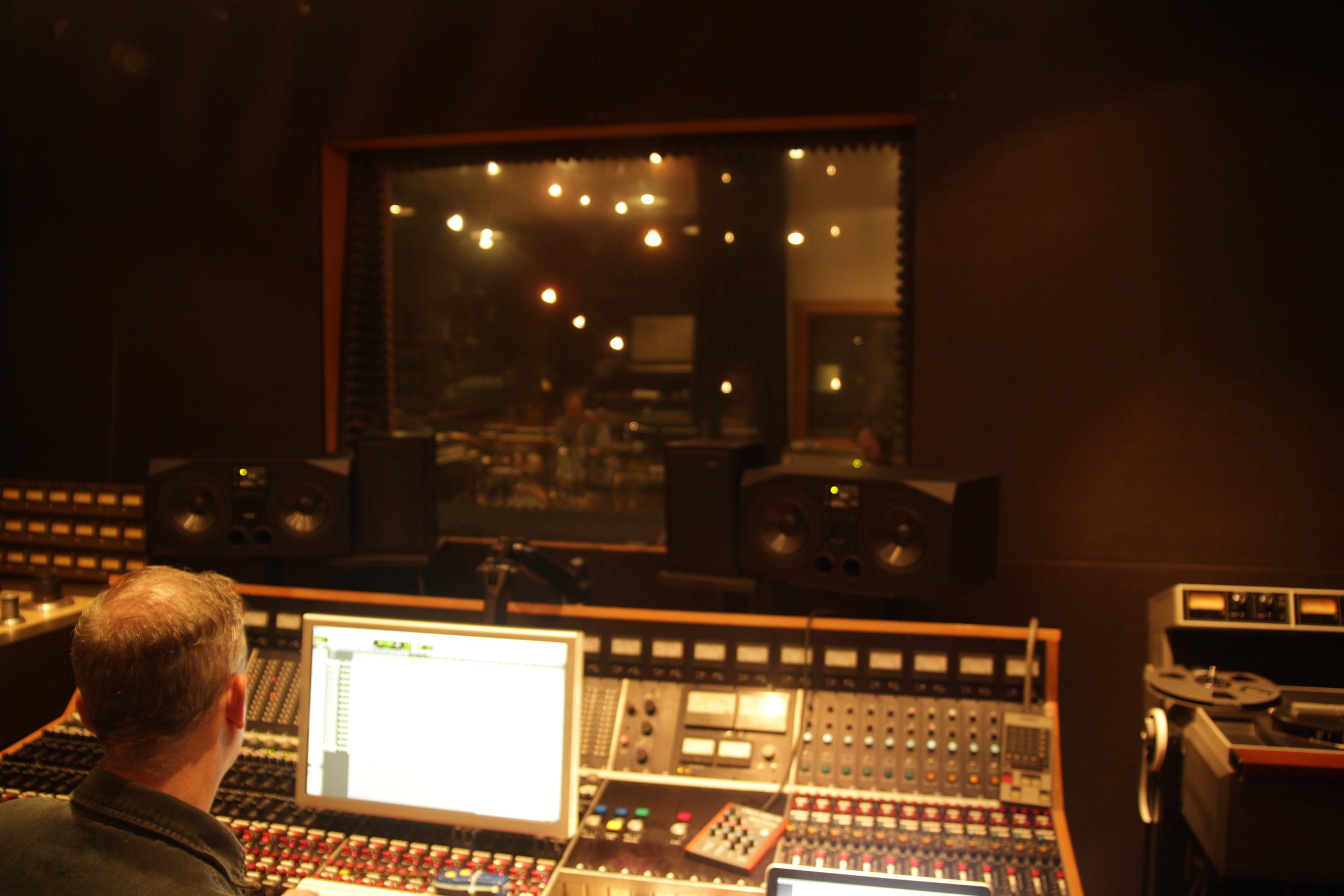 Recording studio control room with engineer at mixing console, large monitor, speakers, and view into live room.