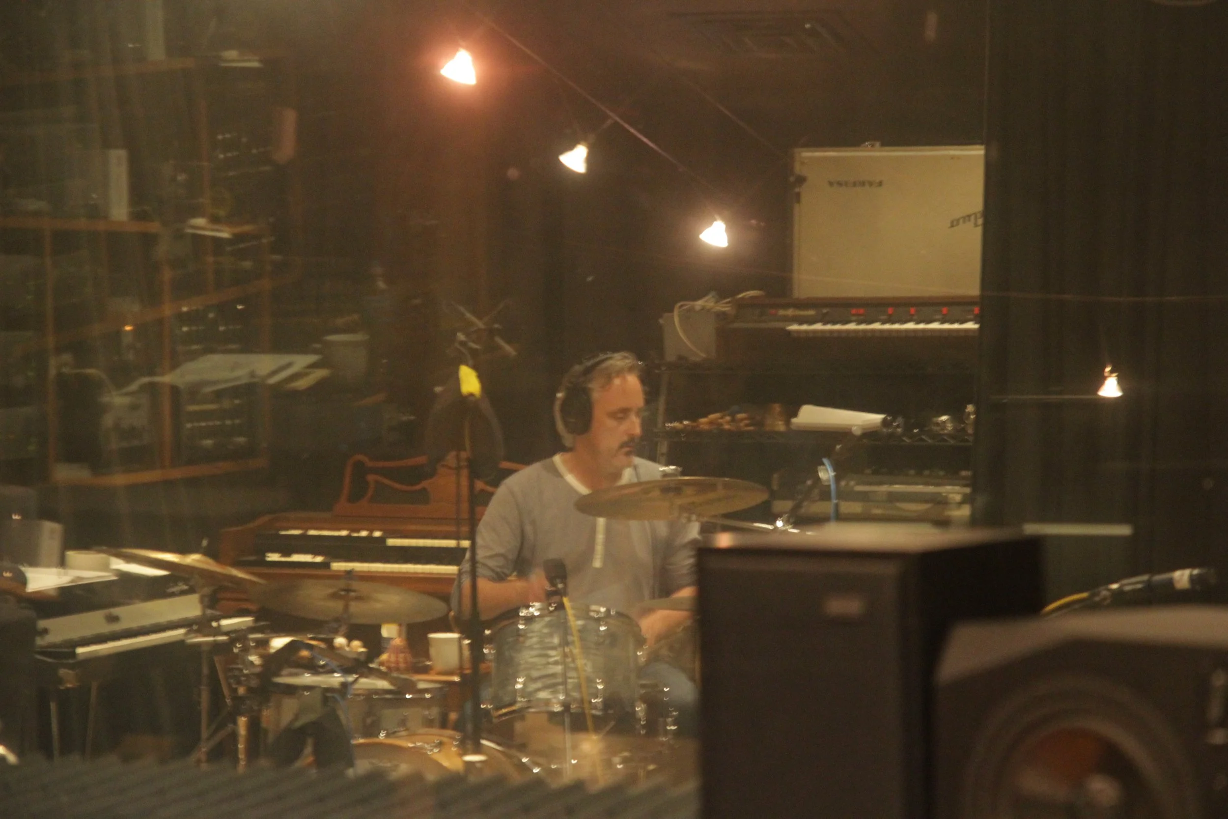 A man wearing headphones playing drums in a recording studio with musical instruments and equipment in the background.
