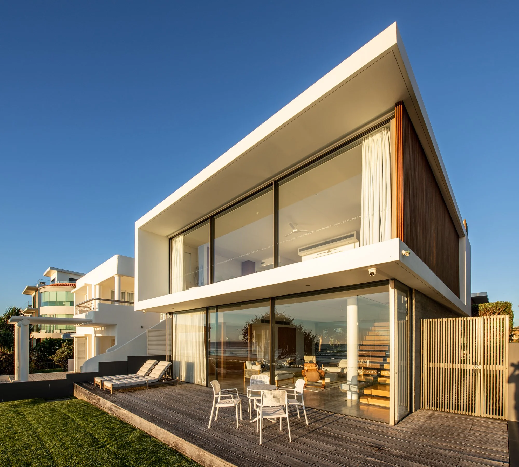 Modern beach house with large glass windows, a wooden deck, and outdoor furniture.