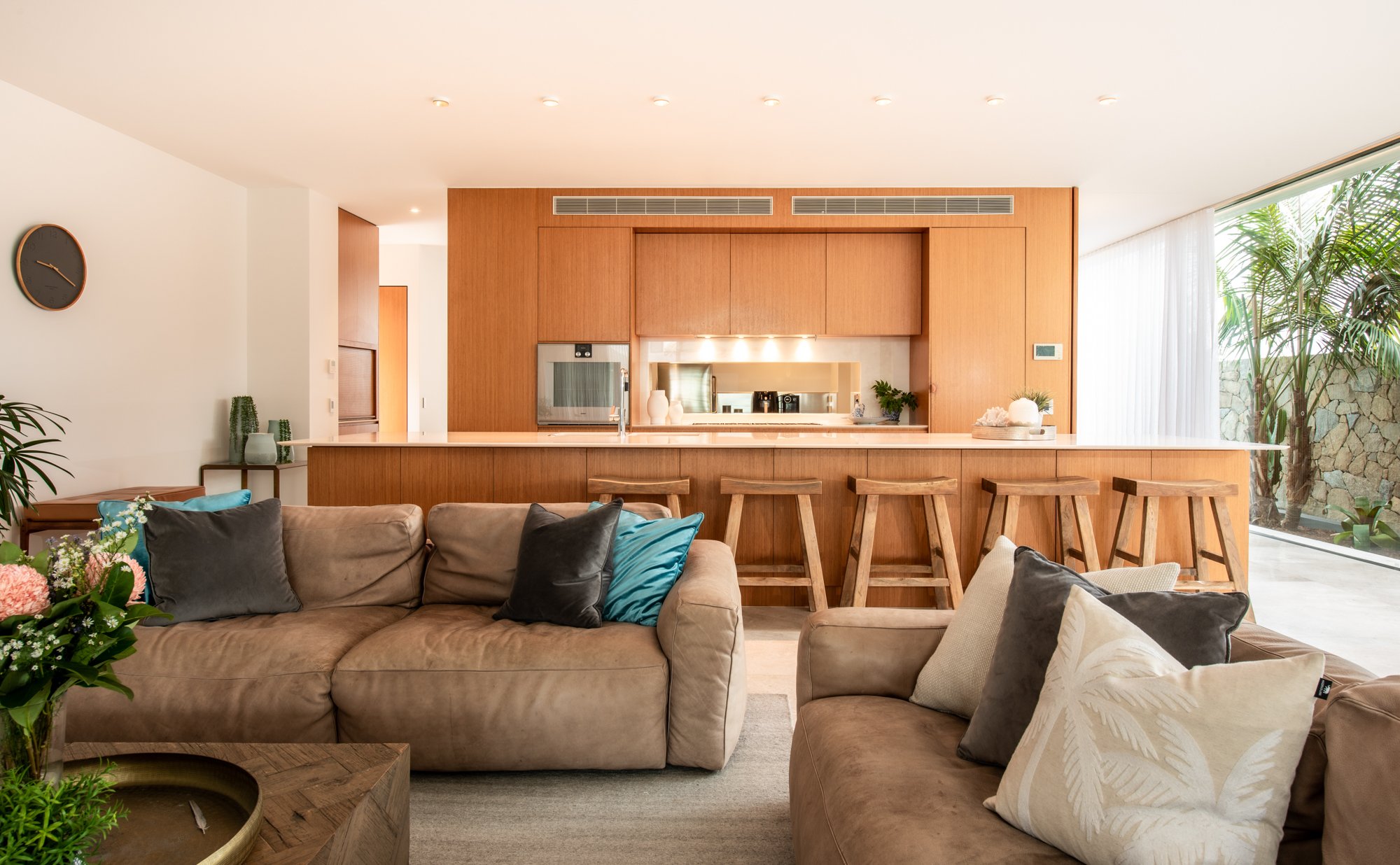 Modern living room with a beige couch and cushions, open kitchen with wooden cabinetry and breakfast bar, stools, large windows, and indoor plants.