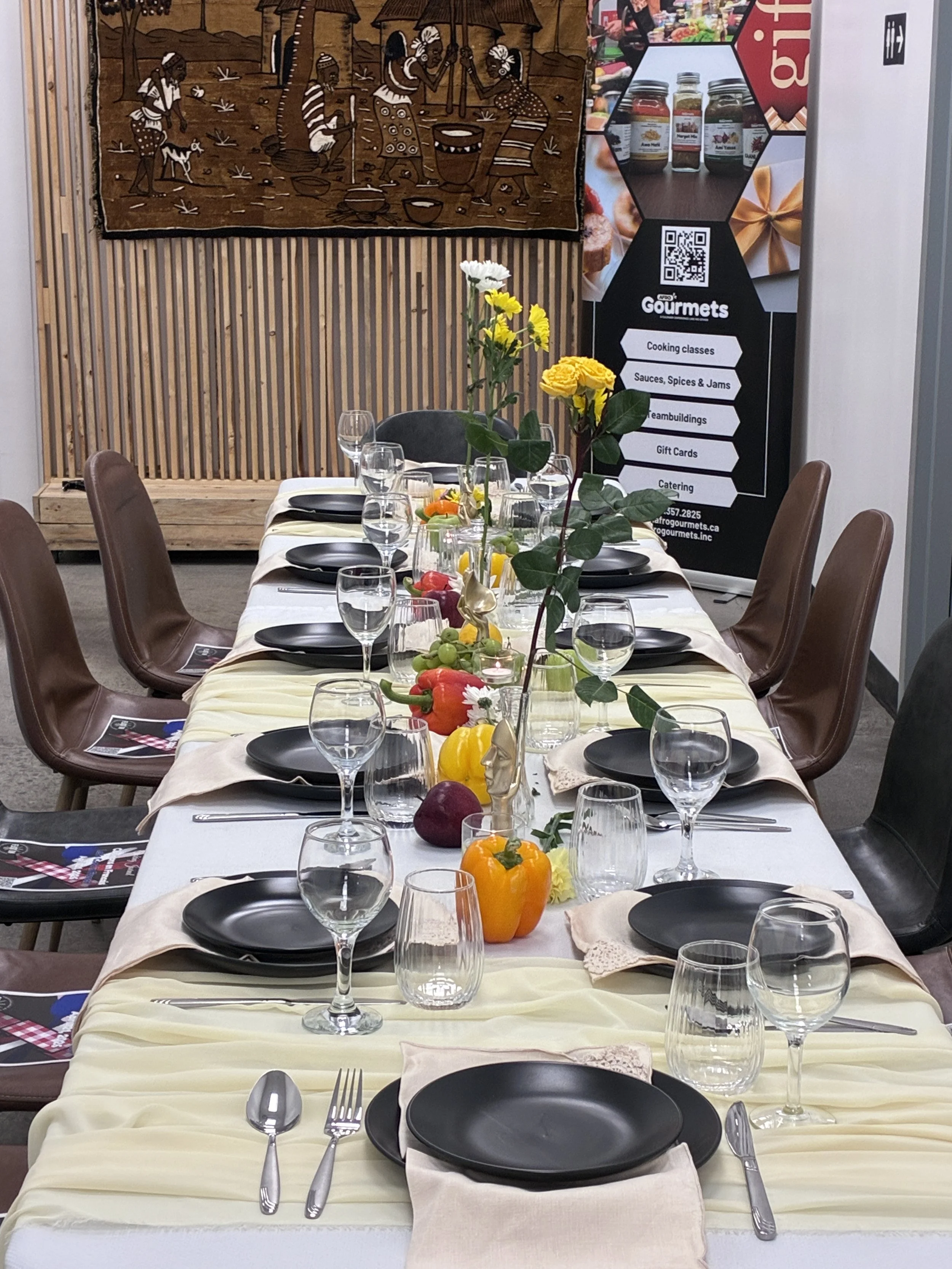 A dining table set for a meal with black plates, silverware, and empty glasses. The table has a yellow tablecloth and a centerpiece of colorful bell peppers and flowers. In the background, there are chairs, a wooden slatted wall, a wall hanging depicting people in traditional African attire, and a promotional stand with various tags and QR codes.