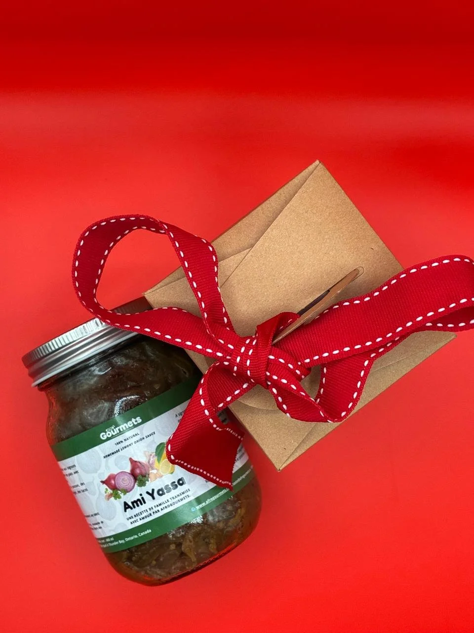 A gift box wrapped with a red ribbon, a jar labeled 'Ami Yassa,' and a small brown paper bag on a red background.