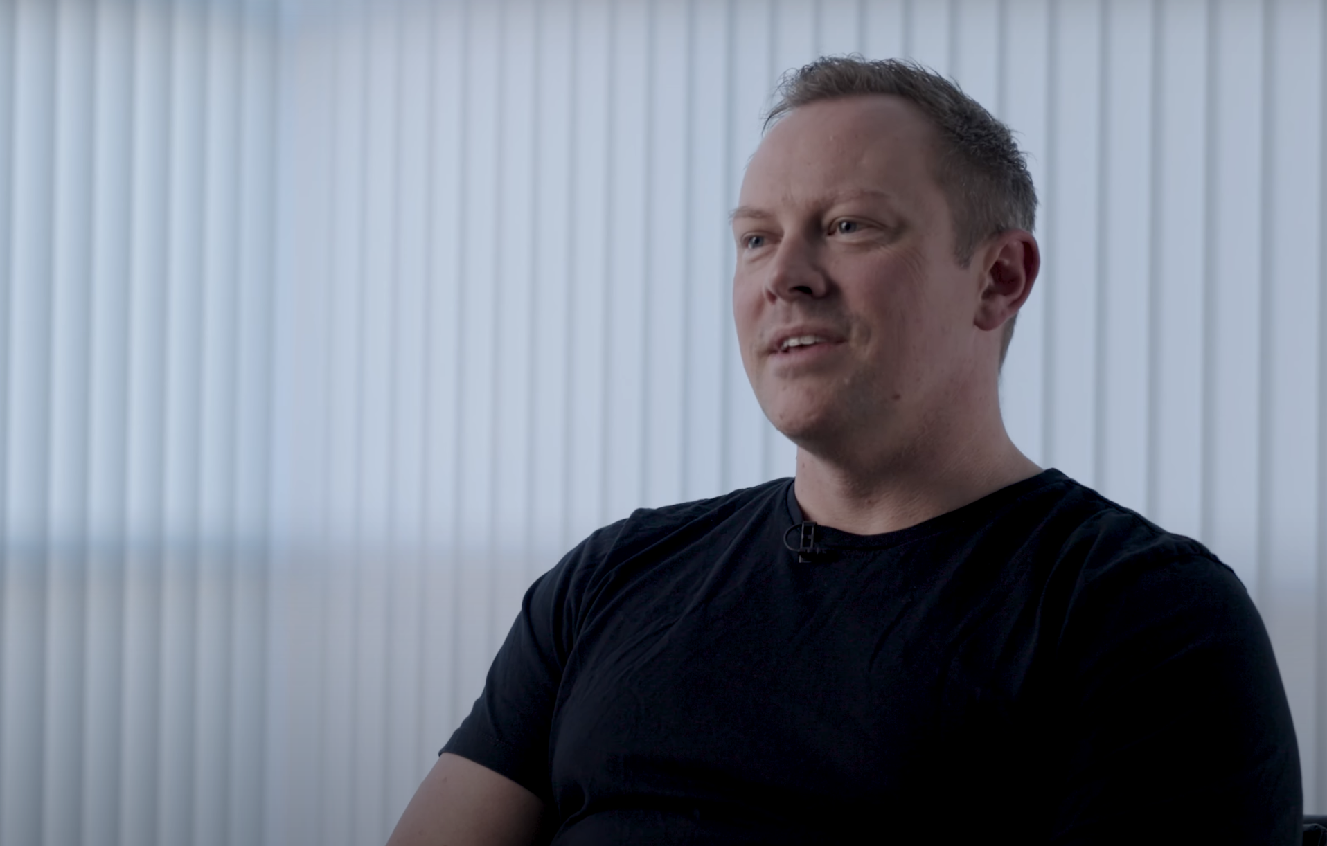 A man with short hair wearing a black shirt, sitting in front of a light-colored, vertically striped background.