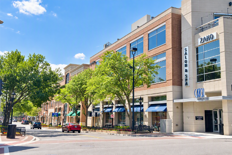 Street view of a commercial area with trees, buildings, and parked cars under a clear blue sky.