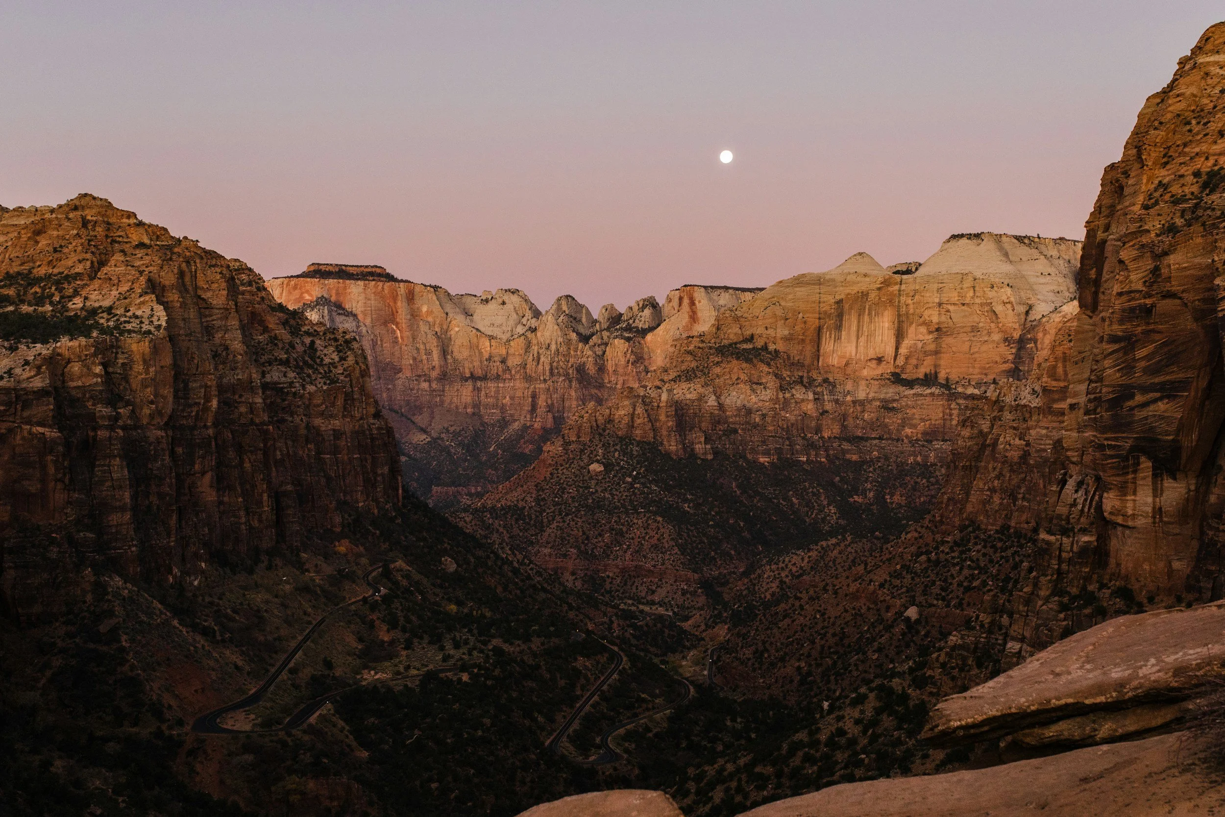 Night view of a canyon with rugged cliffs, winding roads at the bottom, and a visible full moon in the twilight sky.