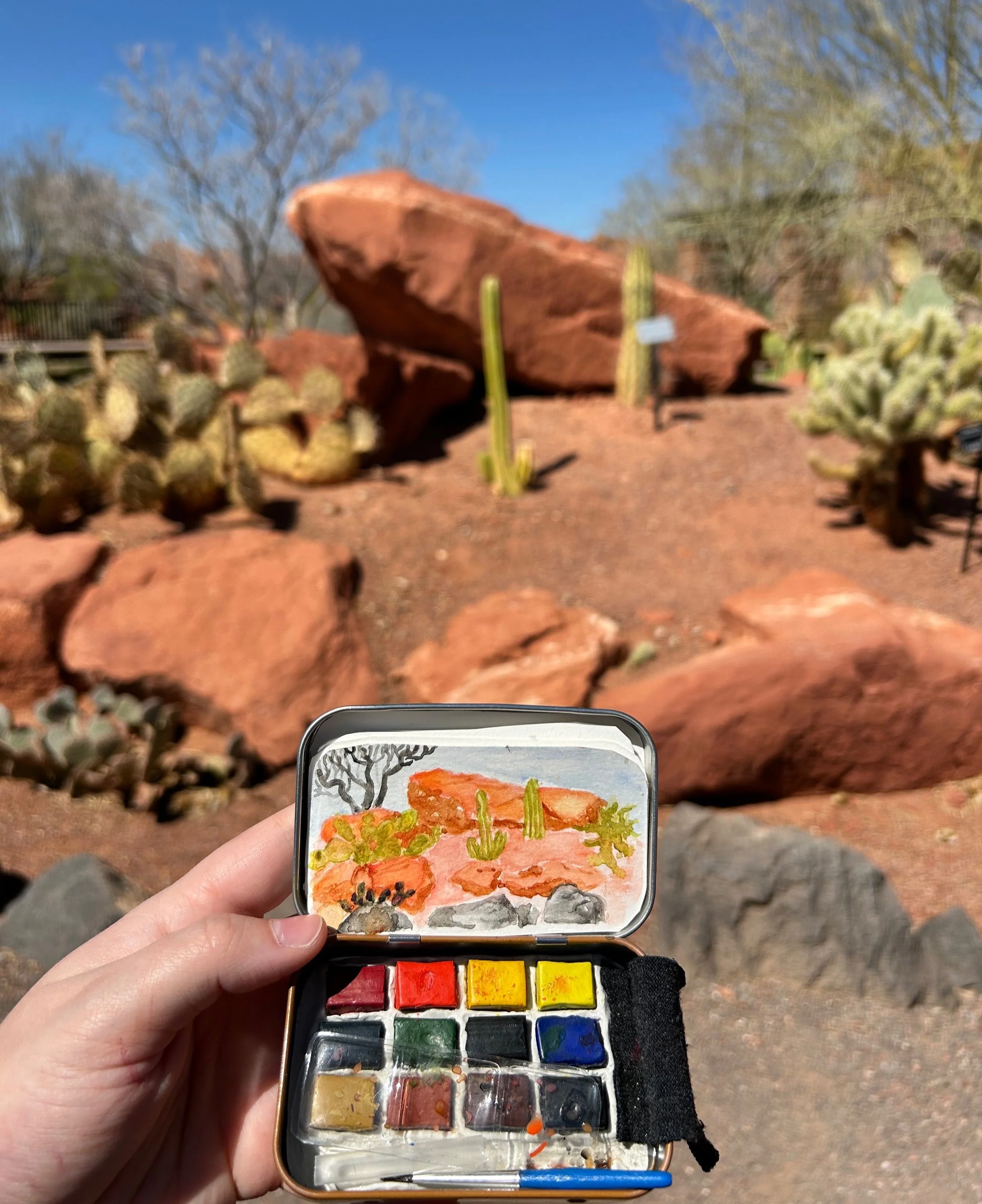 A hand holds a small watercolor paint set and a painting of a desert landscape with rocks, cacti, and dried bushes. The actual desert scene is in the background with large red rocks, cacti, and a clear blue sky.