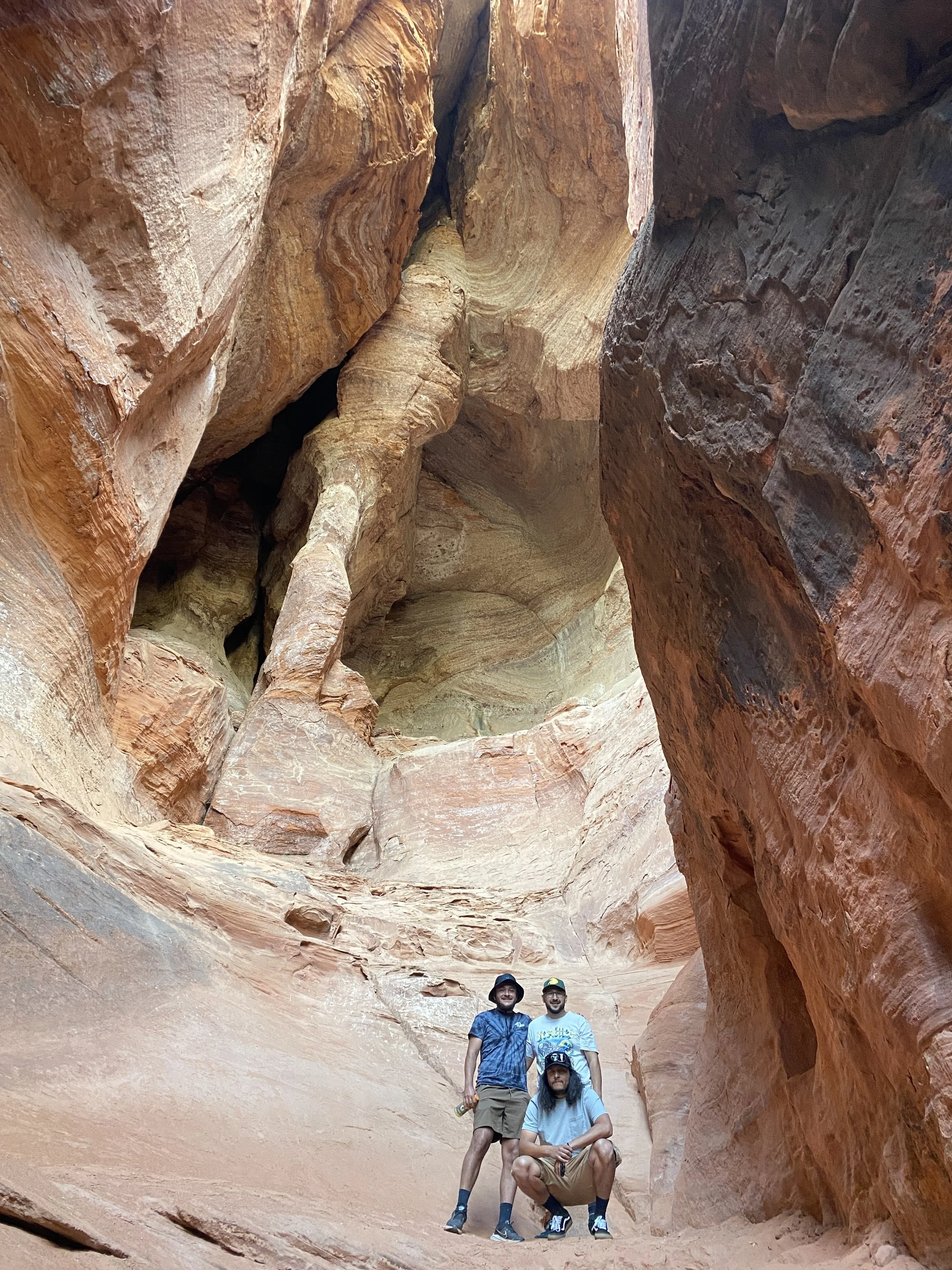 Three men are inside a narrow slot canyon with colorful, layered rock walls, some with curves and overhangs, taking a photo together.