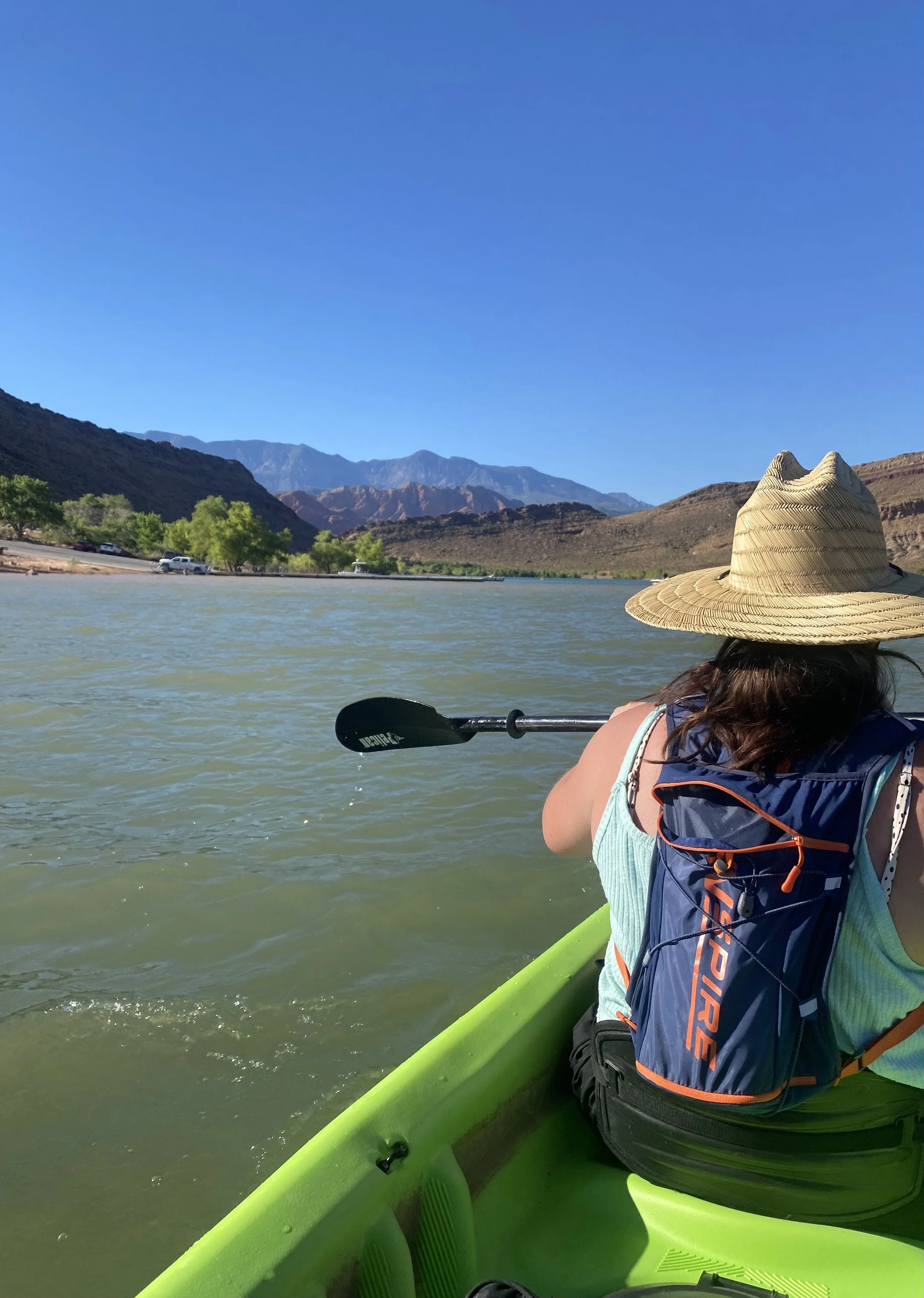 Woman kayaking on a river with mountainous landscape in the background, wearing a straw hat and a blue backpack, under clear blue skies.