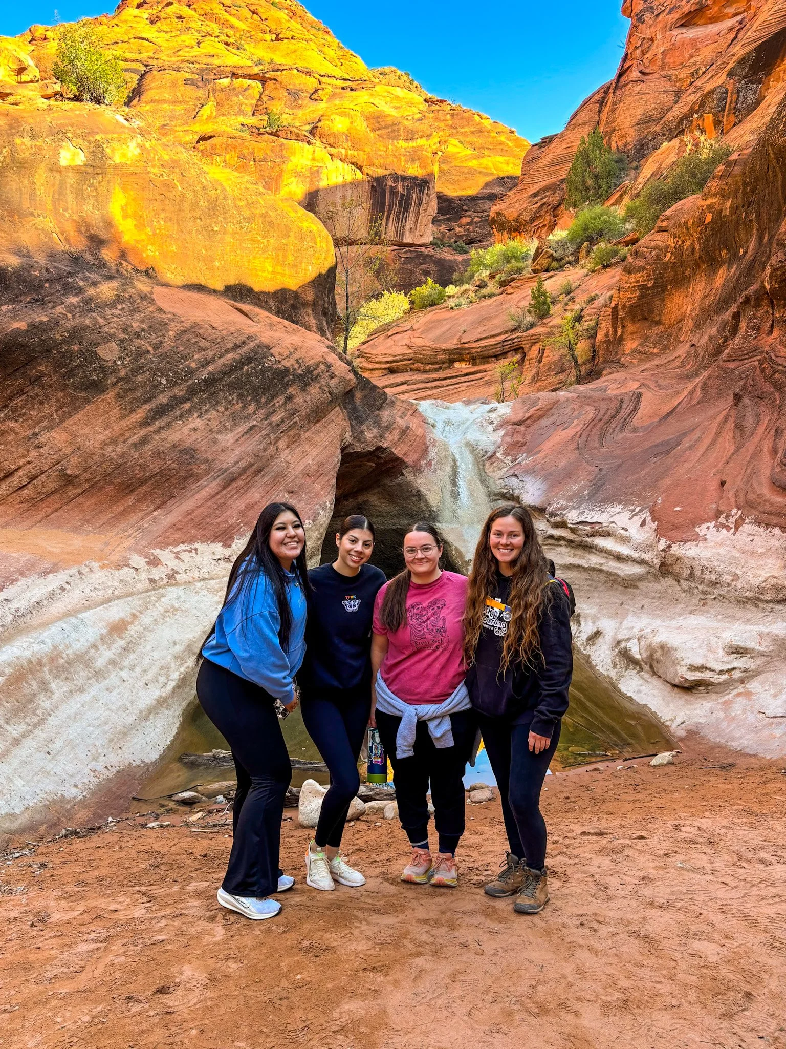Four women standing together in a canyon with red rock walls and a small waterfall behind them, smiling at the camera.