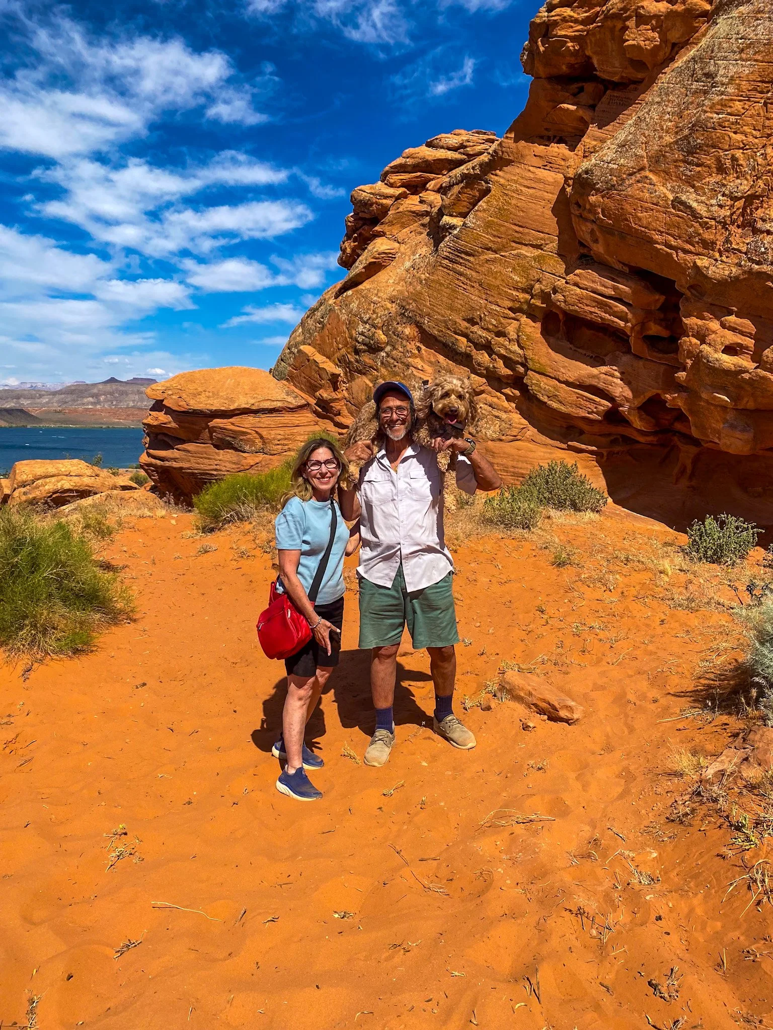 Two people and a dog standing on orange sandy ground with large reddish-brown rock formations in the background and a blue sky with scattered clouds.