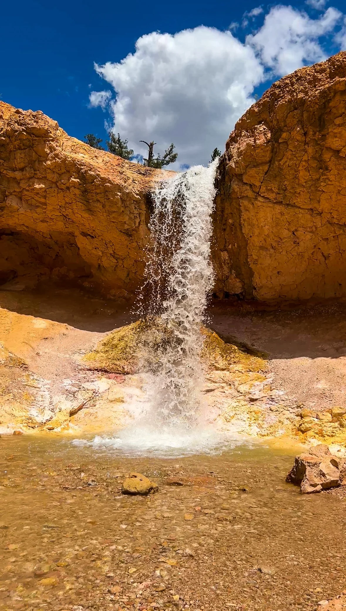 Small waterfall cascading down over orange-brown rocks into a shallow pool, with a bright blue sky and some clouds above