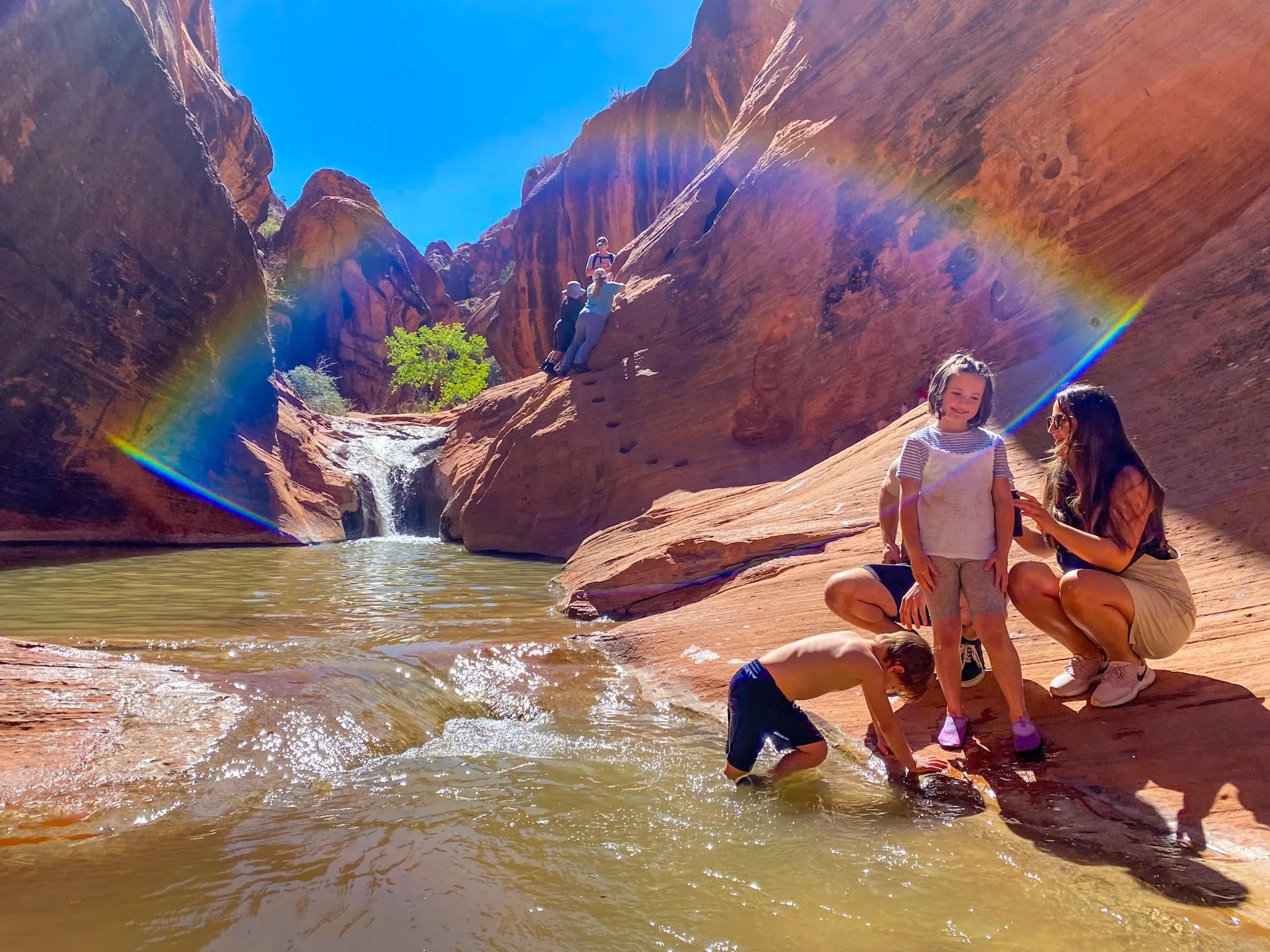 A family enjoying a scenic canyon with a small waterfall, red rock cliffs, and a rainbow lens flare, including two children and two adults.