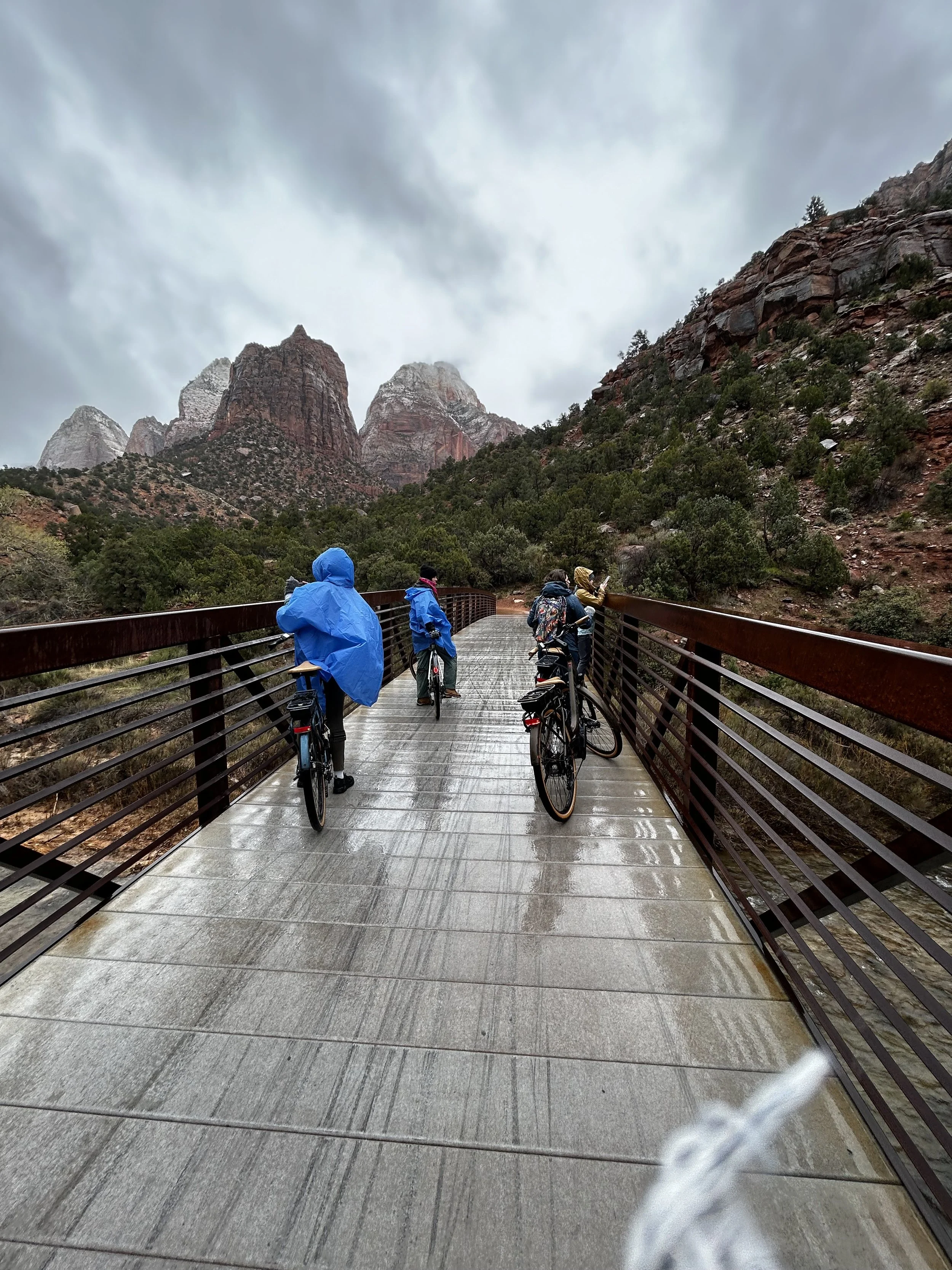 Group of people with bicycles standing on a wooden bridge in a rainy, mountainous landscape with red rock formations and cloudy sky.