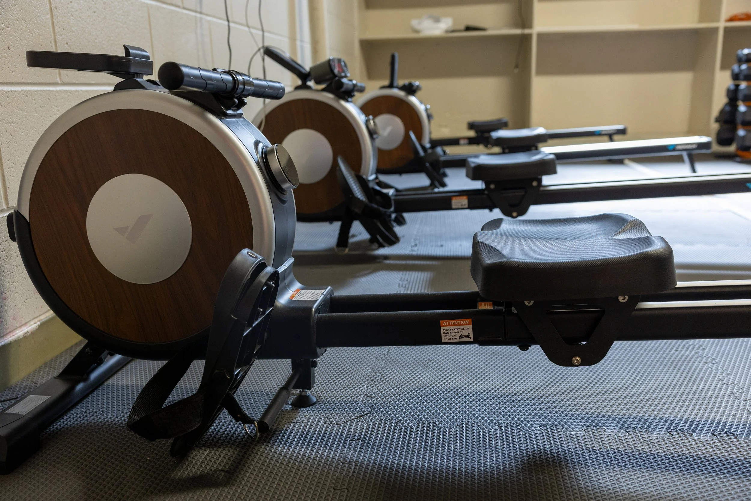 Three rowing machines with black and wood design in a gym setting, positioned on a black rubber floor with a beige wall in the background.