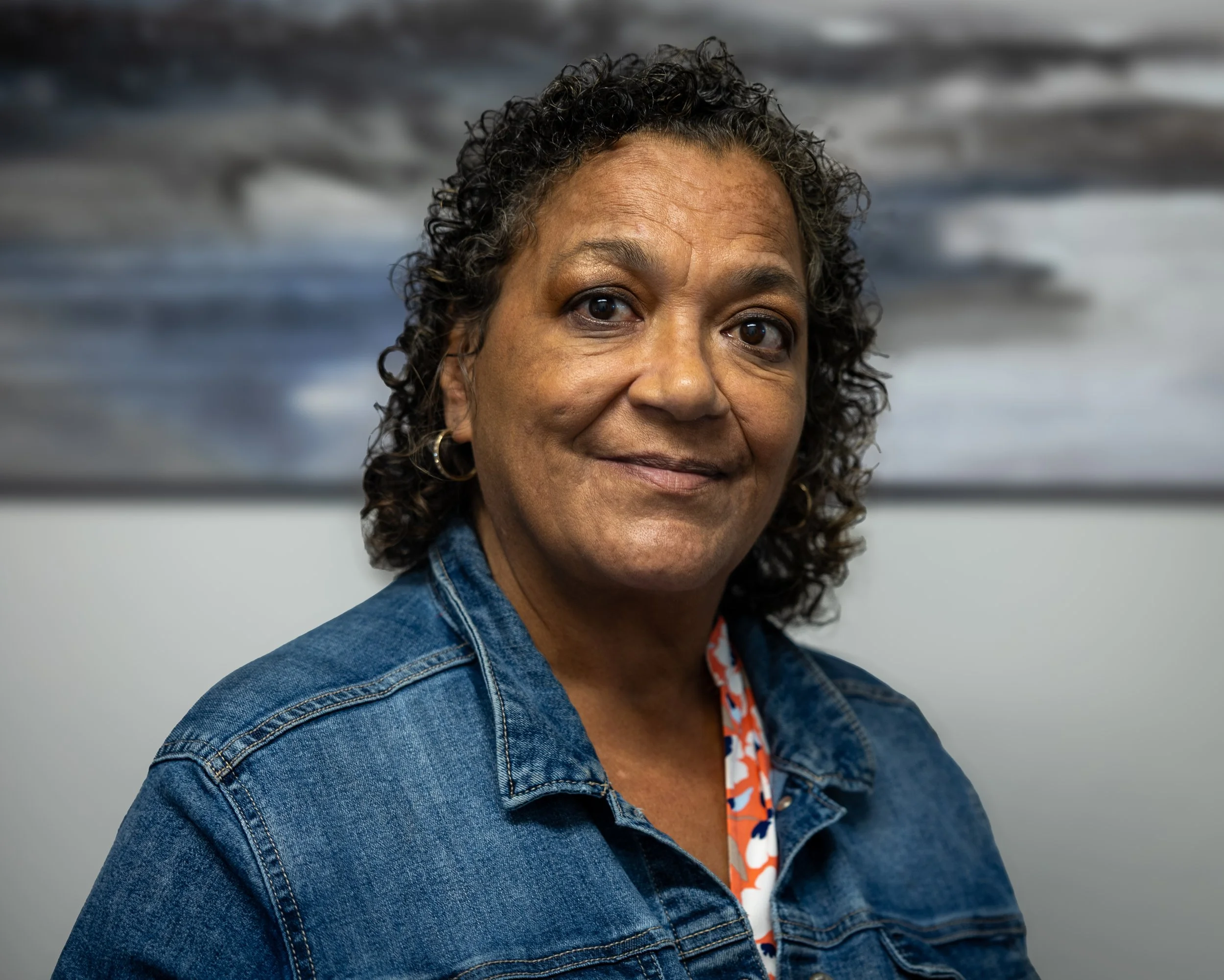 Portrait of a middle-aged woman with black curly hair, wearing a denim jacket and a patterned blouse, standing in front of a background with abstract art.
