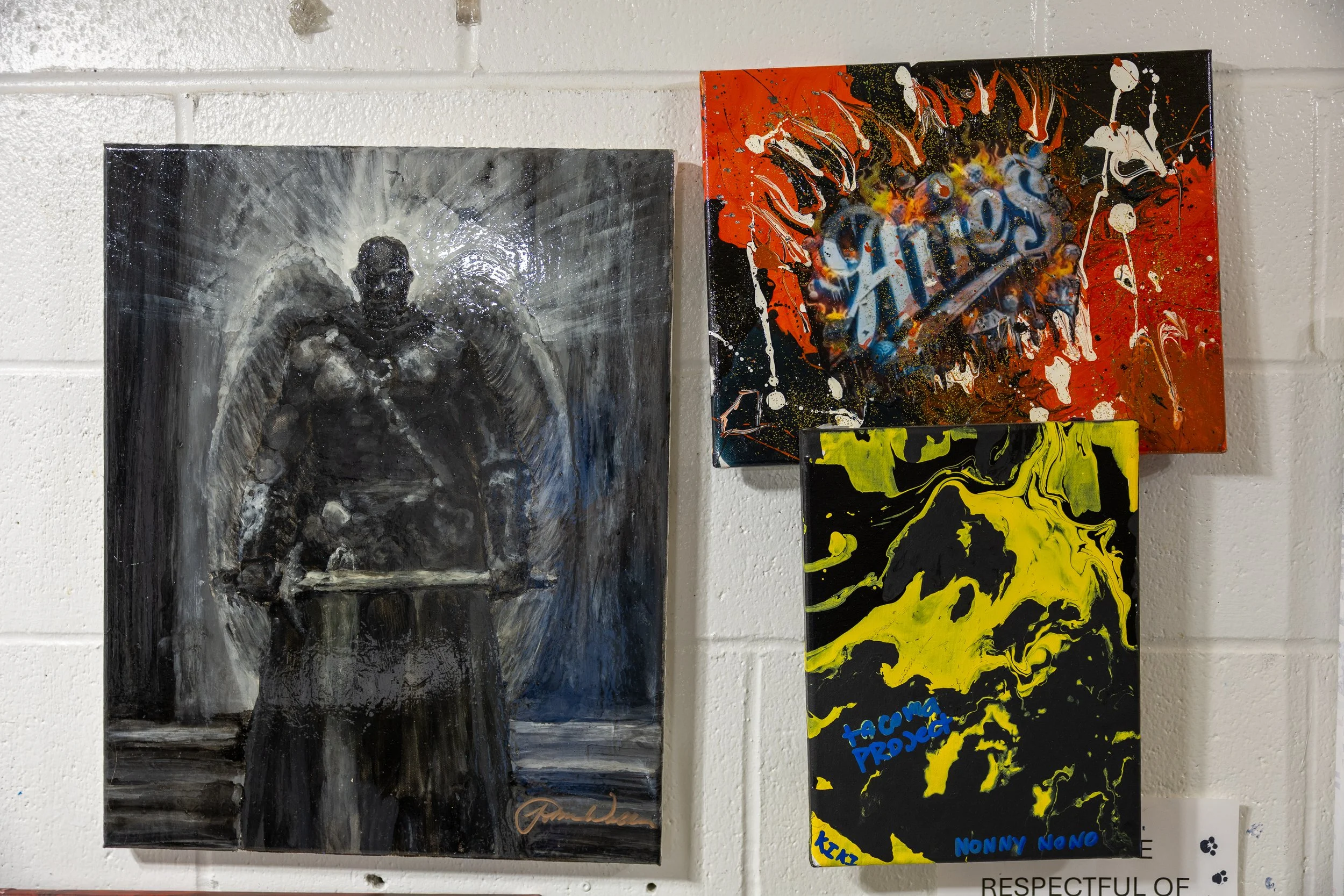 Three abstract paintings hung on a white brick wall, including a monochromatic figure with a halo of light, a colorful splash art piece with the words 'Chaos' and 'Aptos,' and a black and yellow abstract artwork with blue text.