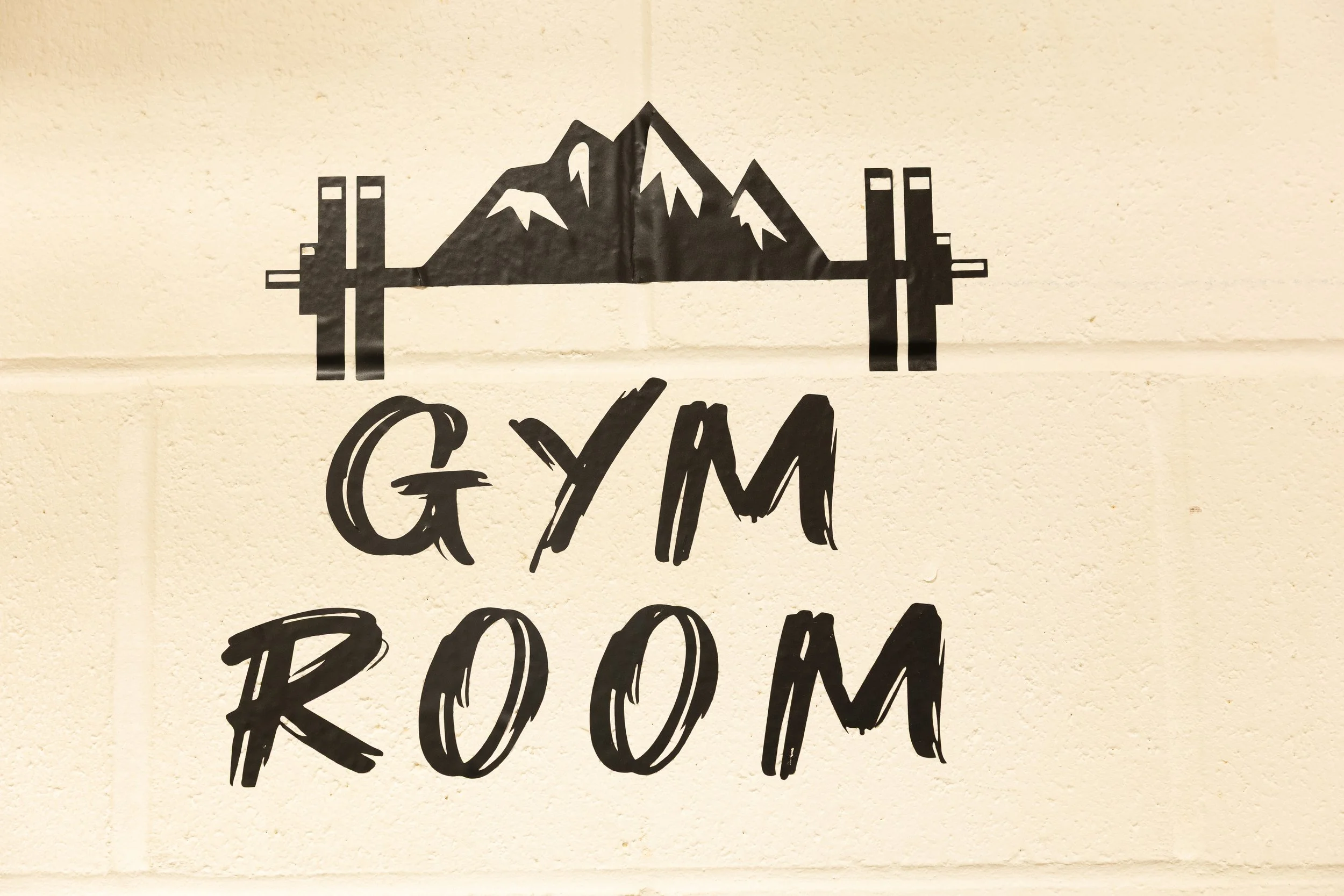 A sign on a beige wall with a mountain and barbell graphic and the words 'GYM ROOM' in bold black lettering.