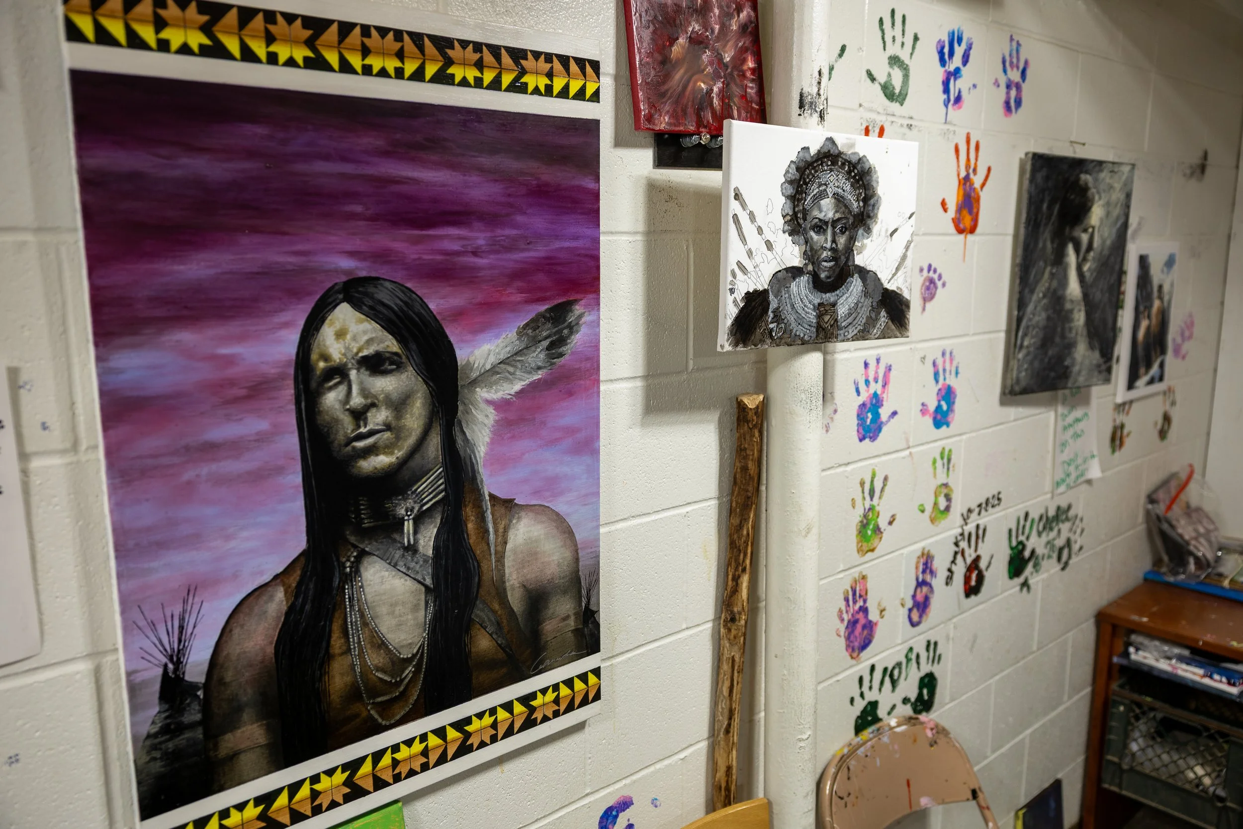 Wall decorated with various colorful and monochrome artworks, including a prominent portrait of a Native American with a feather in her hair, a black-and-white portrait of a woman in traditional jewelry, and several handprint paintings.