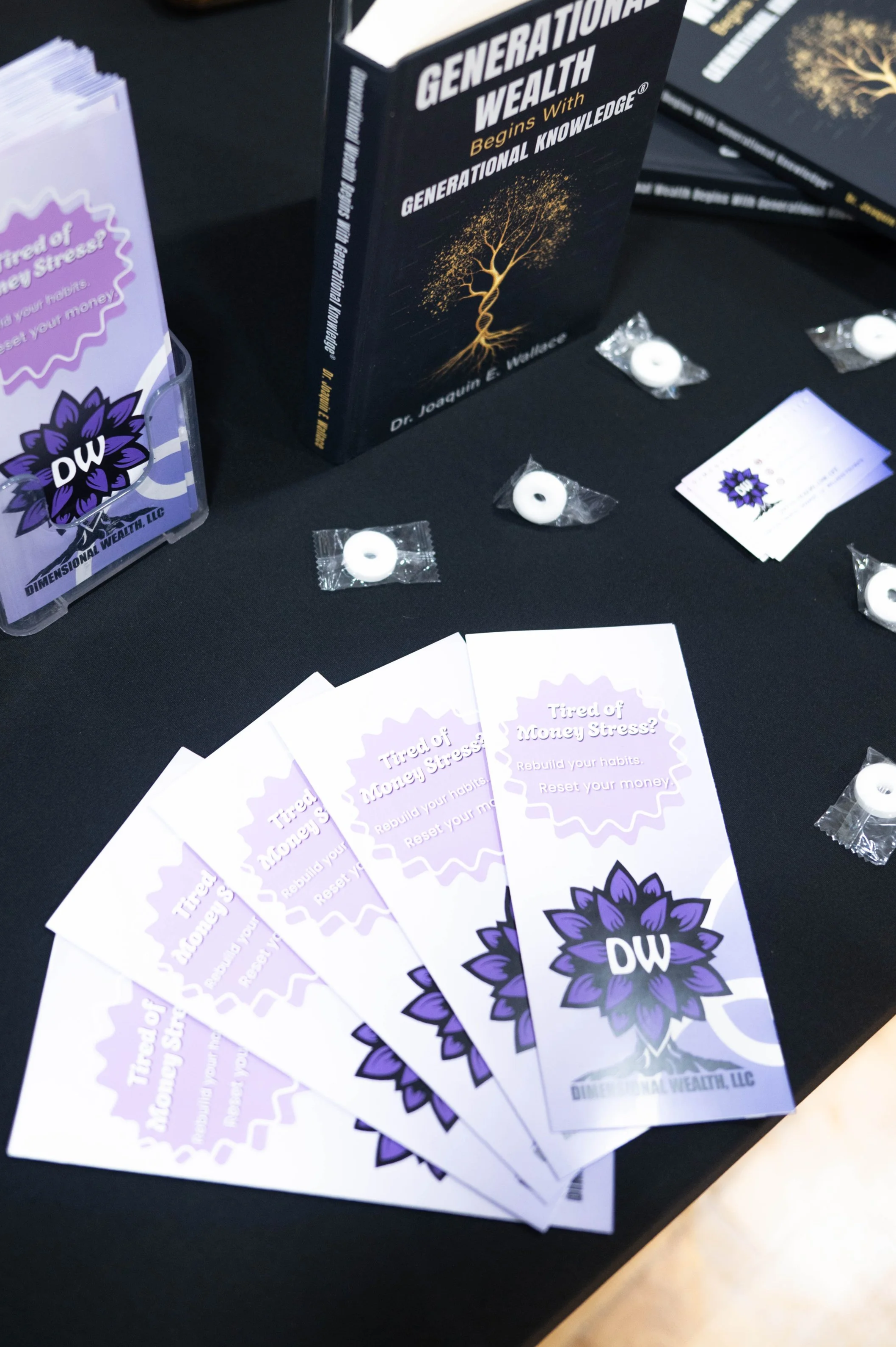 A display table covered in a black tablecloth featuring 'Generational Wealth Begins With Generational Knowledge' books by Dr. Joaquin E. Wallace, surrounded by lavender Dimensional Wealth LLC brochures titled 'Tired of Money Stress?'.