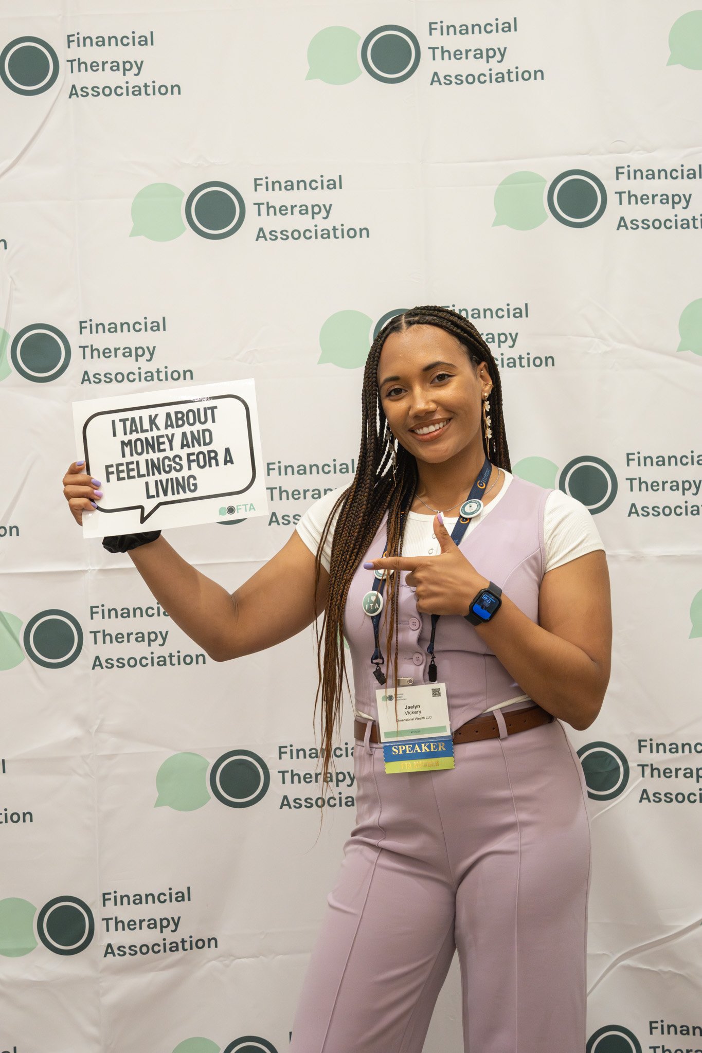 Jaelyn Vickery smiling at the Financial Therapy Association conference holding a speech bubble sign that says "I talk about money and feelings for a living" and pointing to her speaker badge.