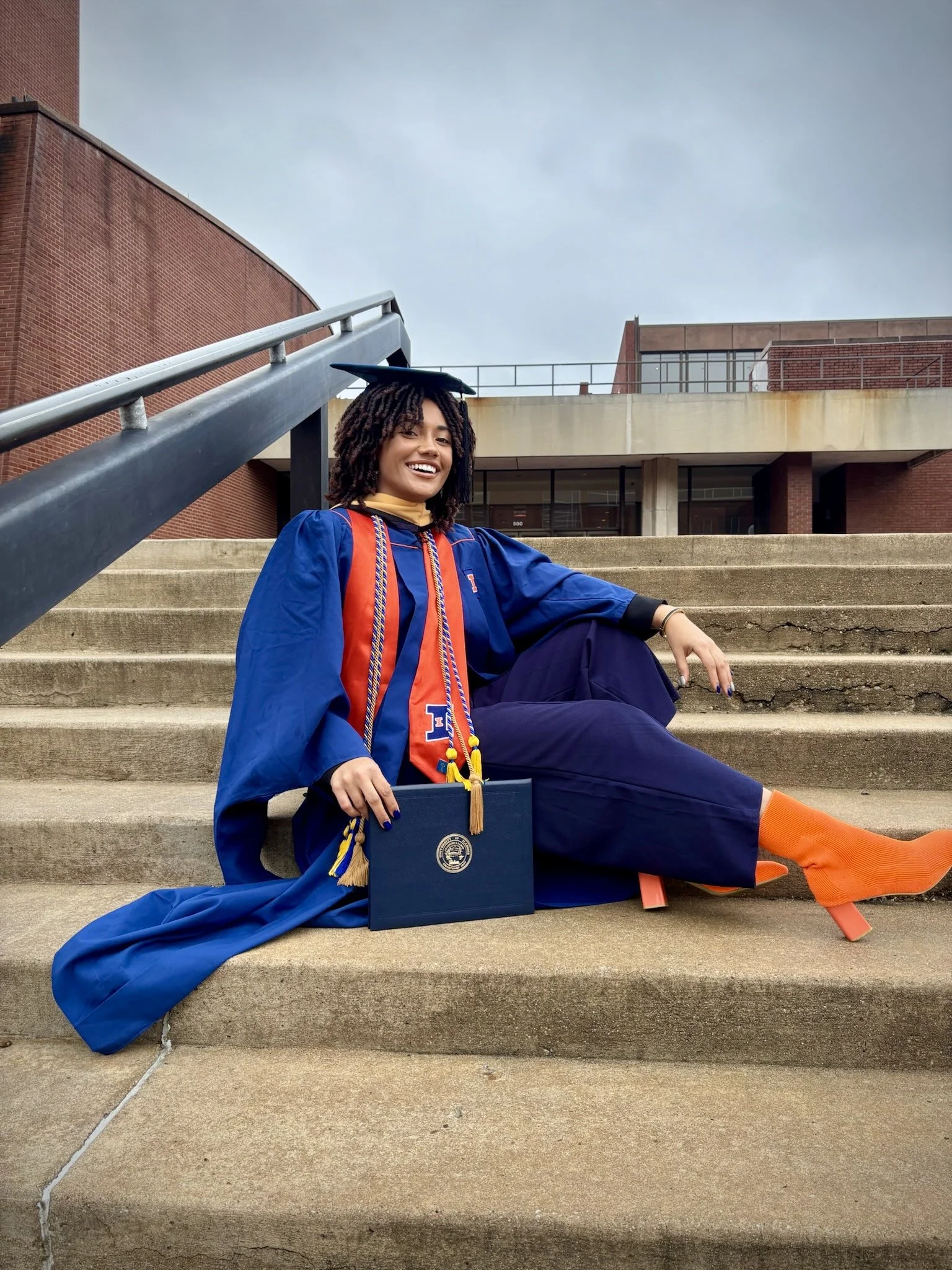 Jaelyn Vickery sitting on concrete steps in her blue and orange graduation gown holding her Master of Social Work diploma folder.