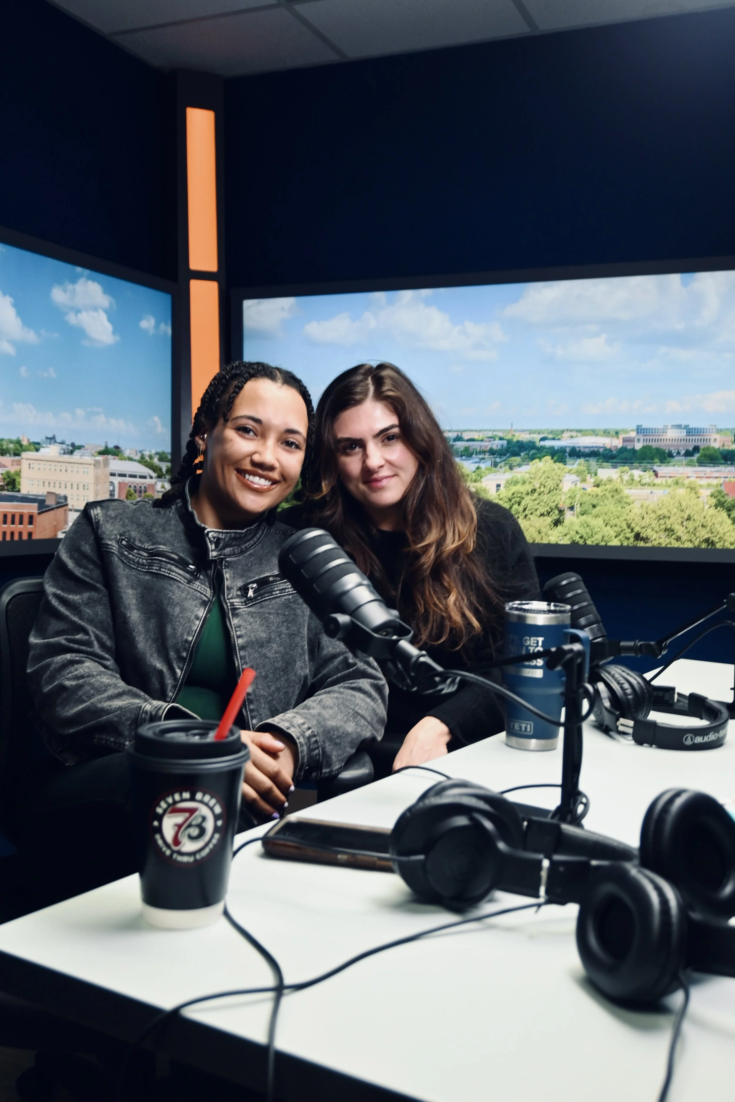 Close-up of Jaelyn Vickery and Grace Ganley smiling together behind a podcast desk at WCIA, featuring a blue Yeti travel mug that reads 'Let's Get Down to Business.'