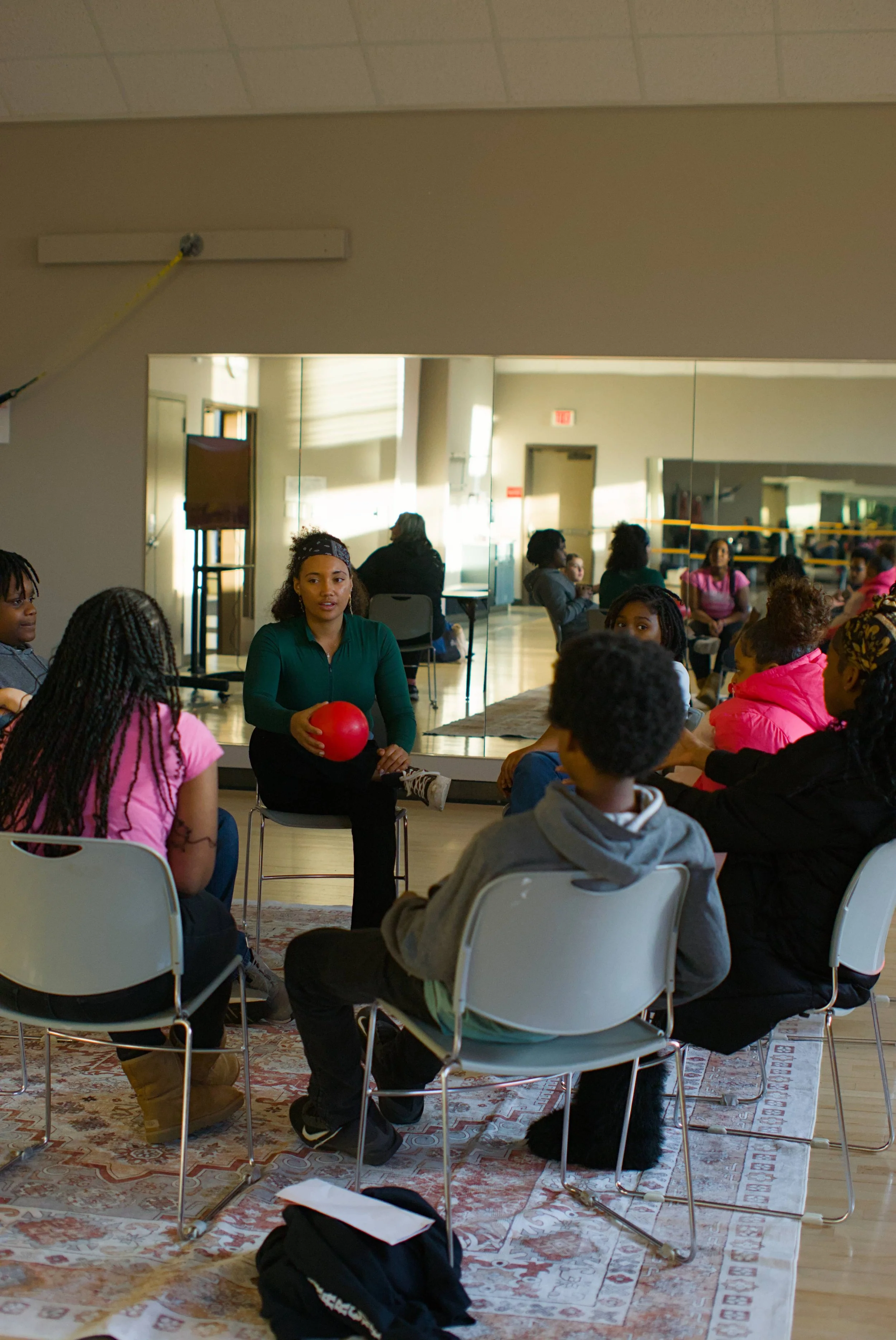 Woman sitting in a circle with teenagers holding a red ball speaking with the participants about financial literacy and wellness.