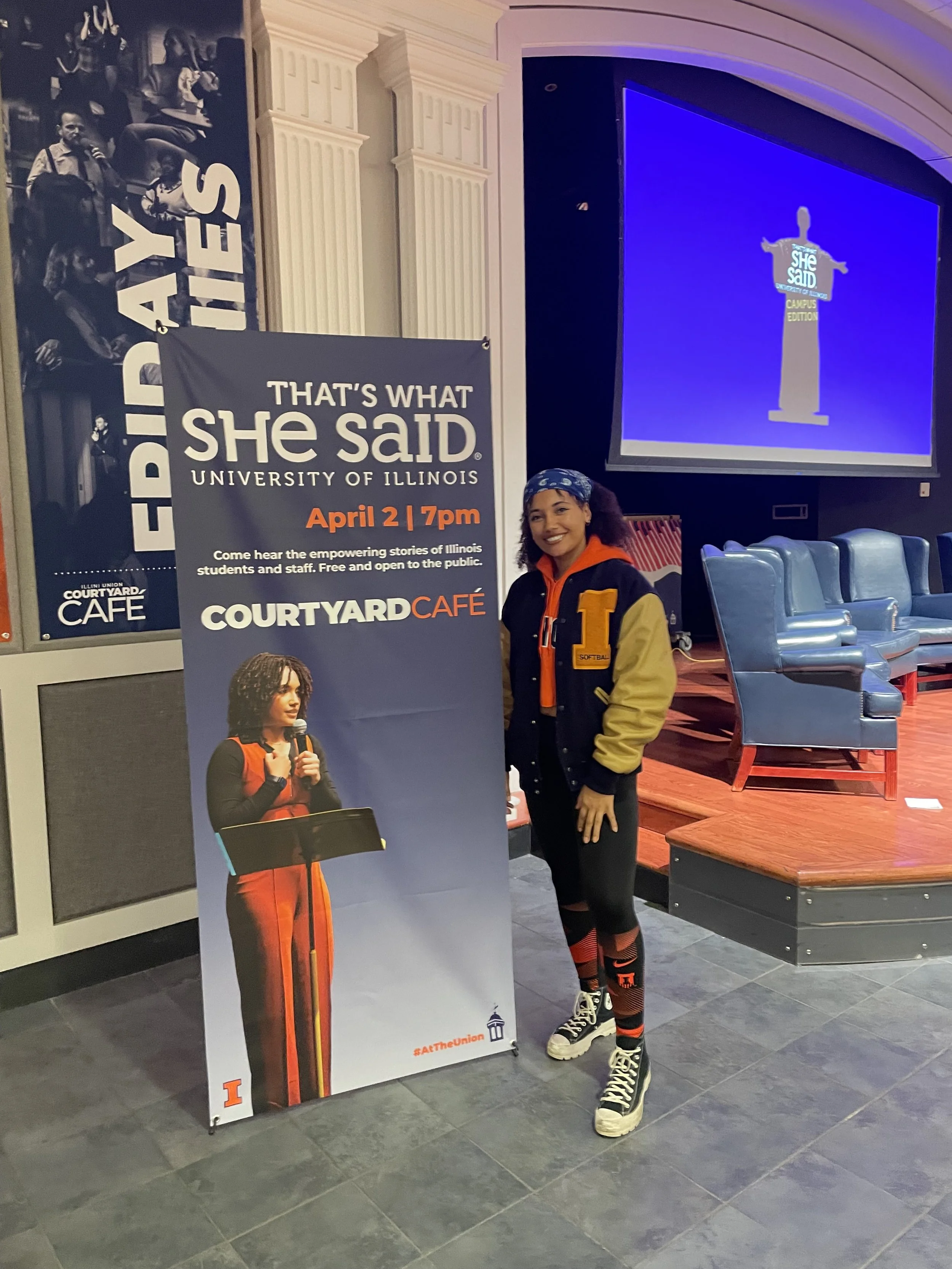 Event emcee standing next to That’s What She Said Campus Edition event banner at the University of Illinois Courtyard Café before a women’s storytelling program.