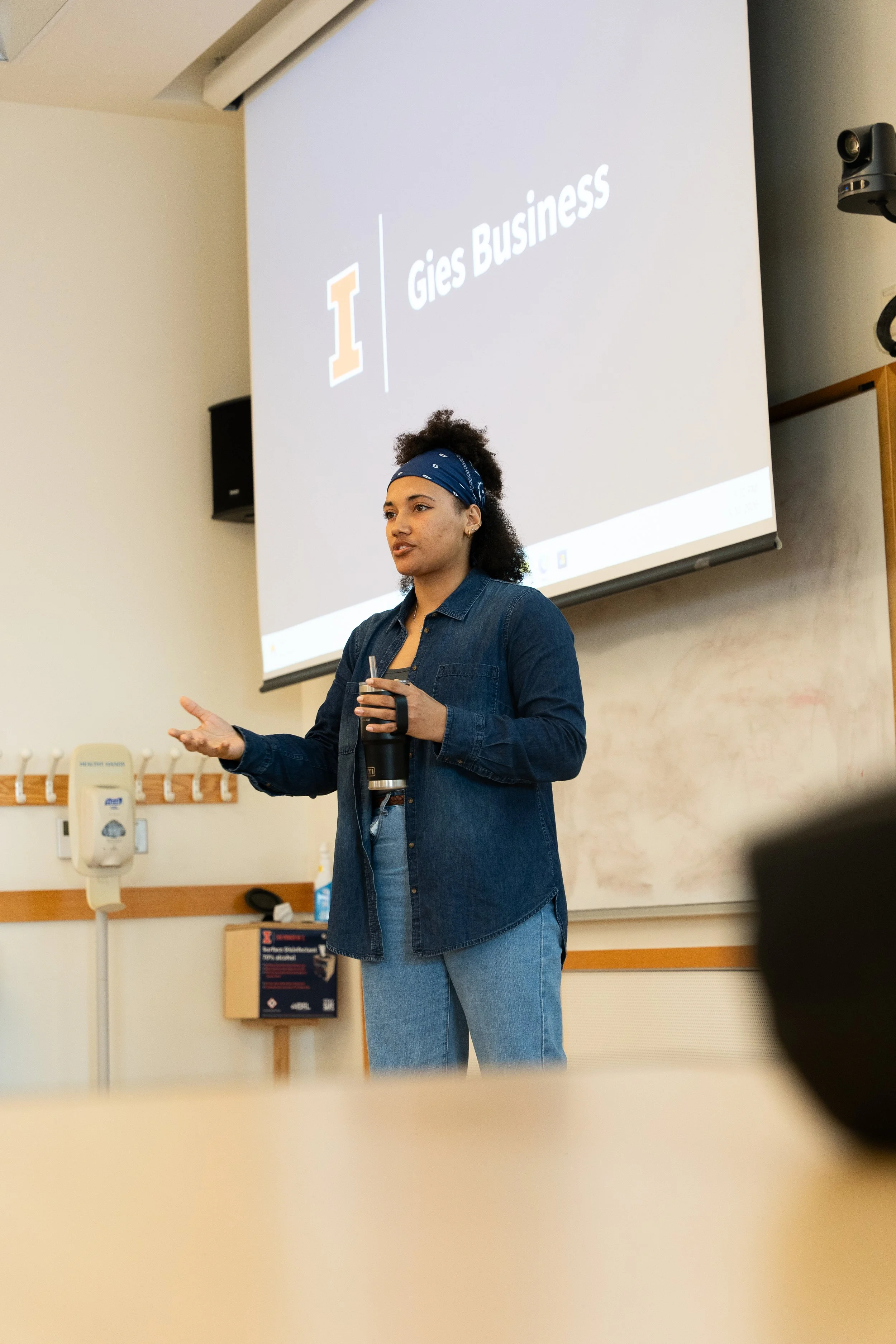 A female financial therapist and coach wearing a blue bandana and denim shirt presenting at Gies College of Business, gesturing during a workshop with a "Gies Business" logo projected behind her.