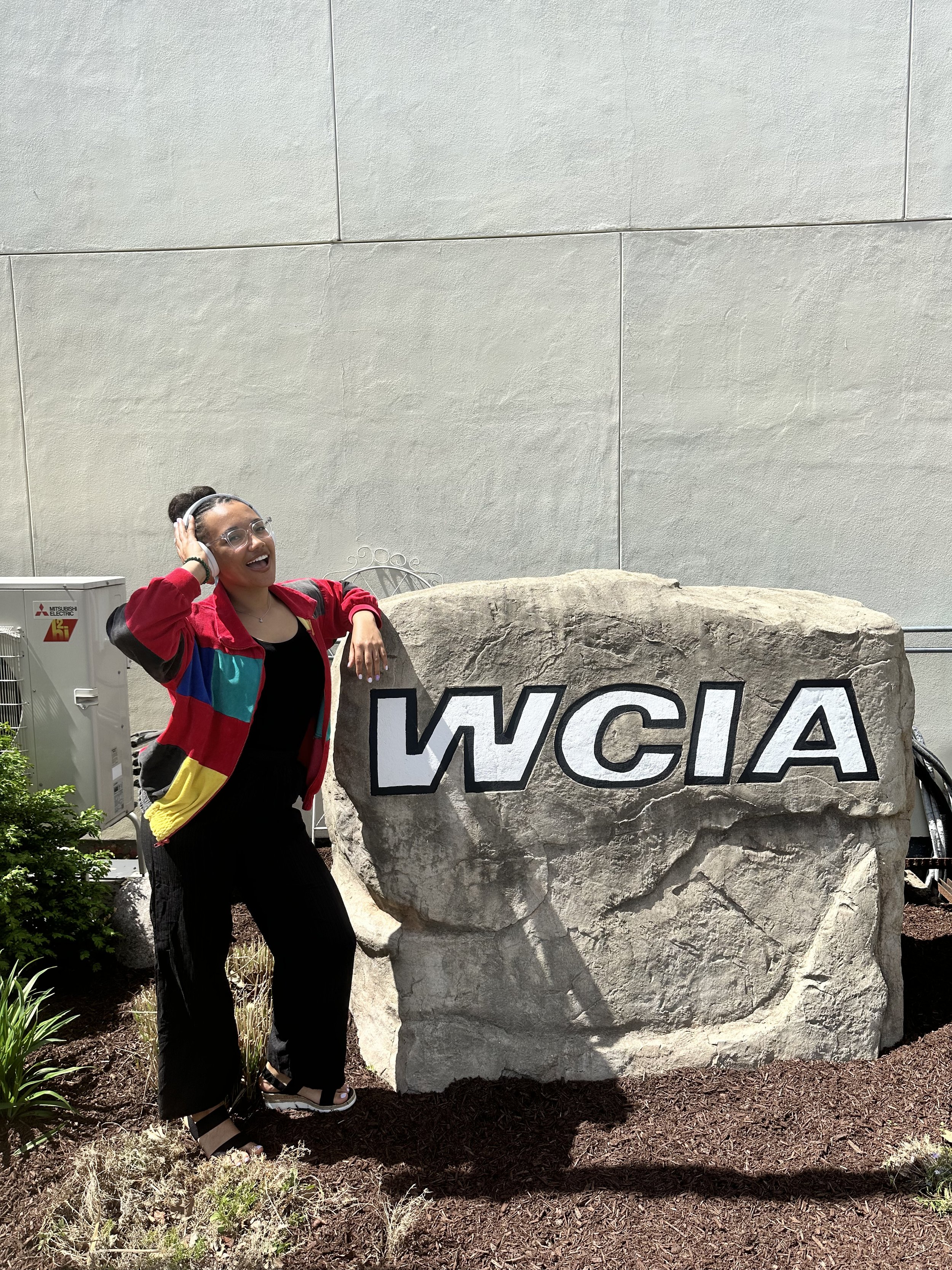 Standing outside the WCIA studio, a smiling woman named JaeVic poses beside a large engraved station sign, capturing a moment that represents the growth of Finance, Fitness & Finesse (F3) from a personal project to a professional media partnership.