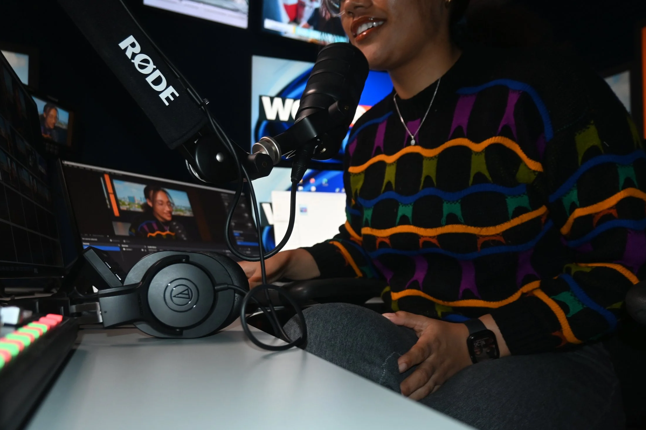 Podcast host JaeVic speaking into a studio microphone during a Finance, Fitness & Finesse recording session, surrounded by screens, audio equipment, and colorful studio lighting while creating content on finance, wellness, and mindful money habits.