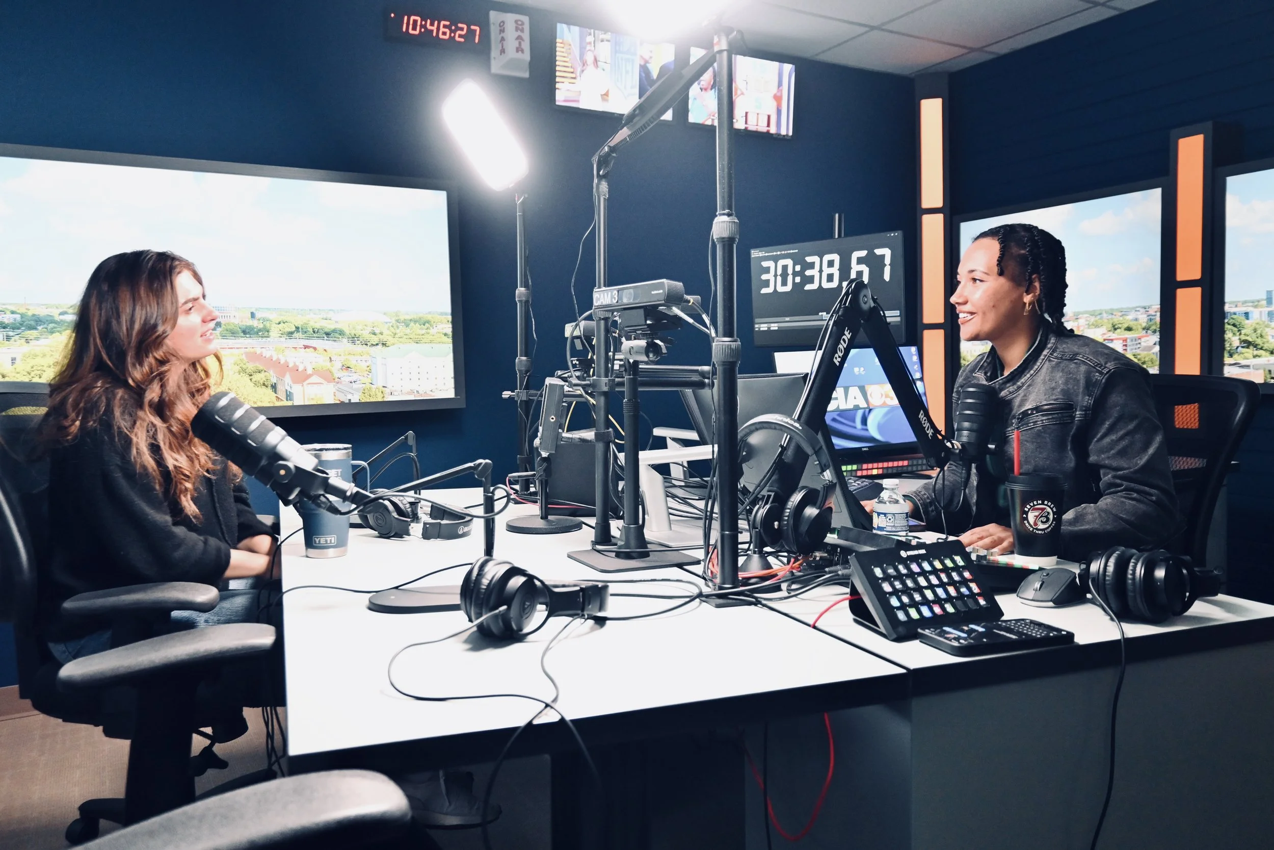 A wide shot of the Finance Fitness Finesse podcast studio set where host Jaelyn Vickery interviews Grace Ganley of In Good Company at a desk equipped with microphones, mixing boards, and digital monitors.
