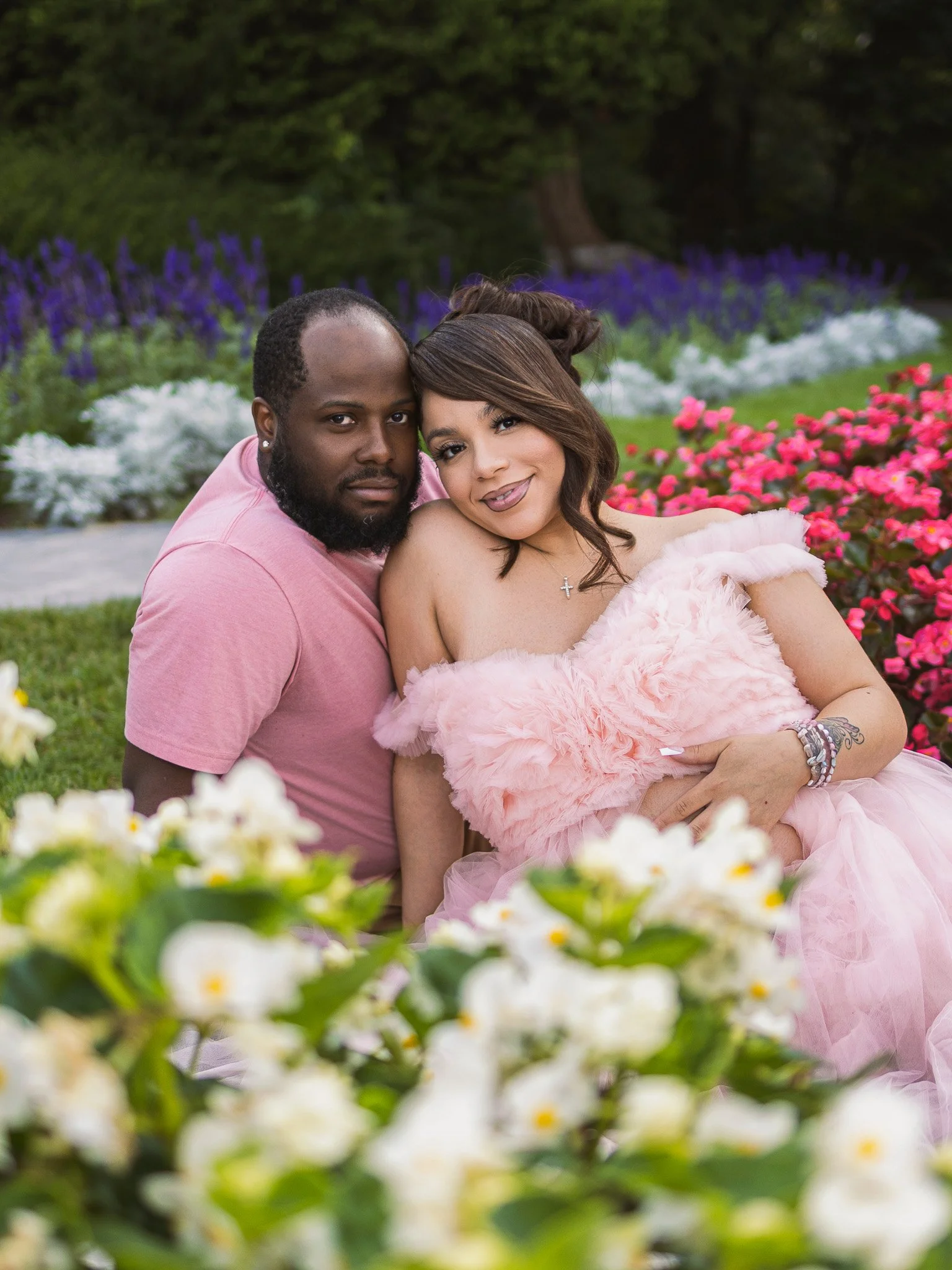 A couple sitting close together outdoors among colorful flowers and greenery, smiling at the camera.