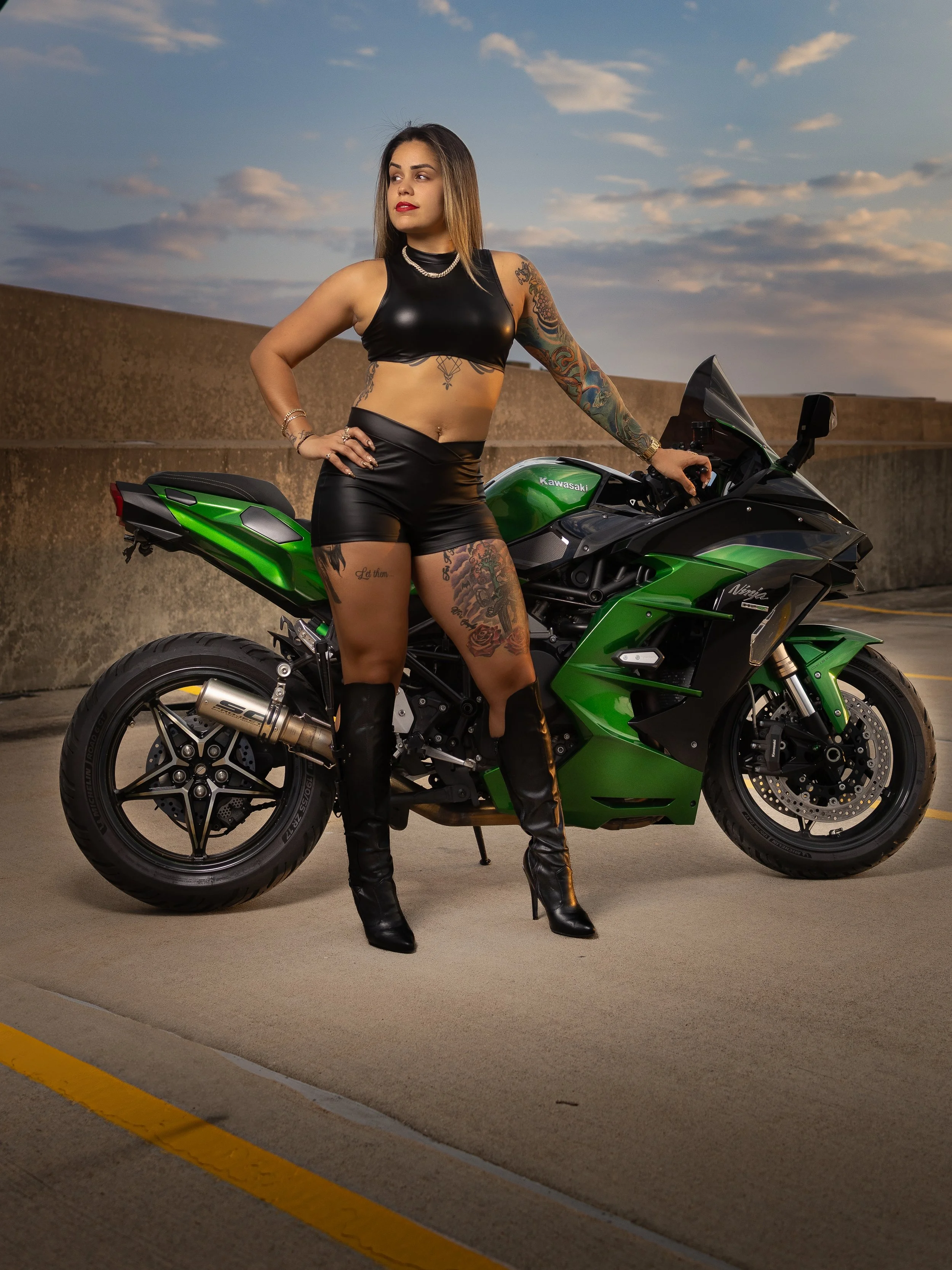 A woman with tattoos standing next to a green Kawasaki motorcycle in a parking lot at dusk.