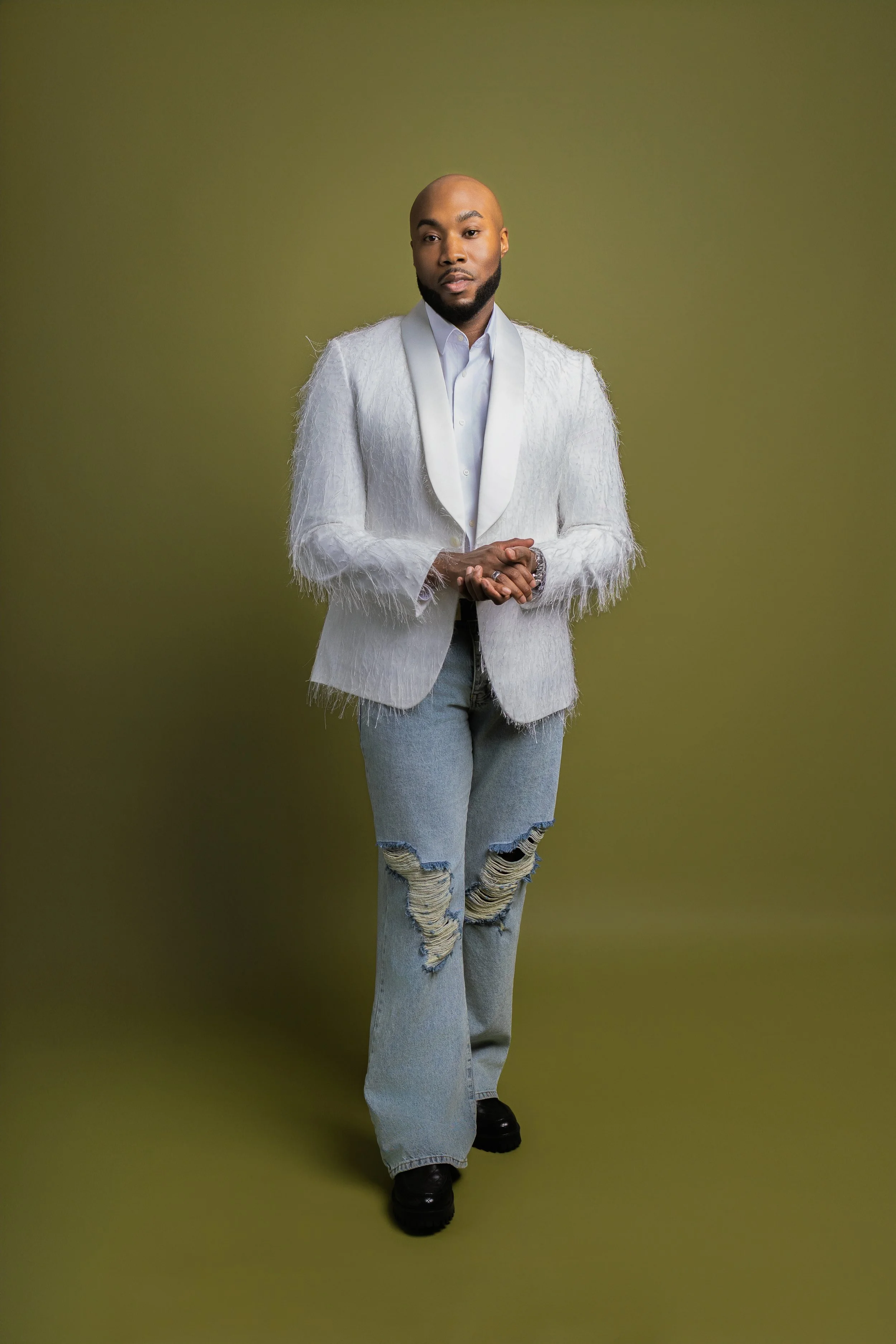 A man with a shaved head, beard, and dark skin wearing a white textured blazer with satin lapel, white shirt, ripped gray jeans, and black shoes, standing against a plain olive green background.