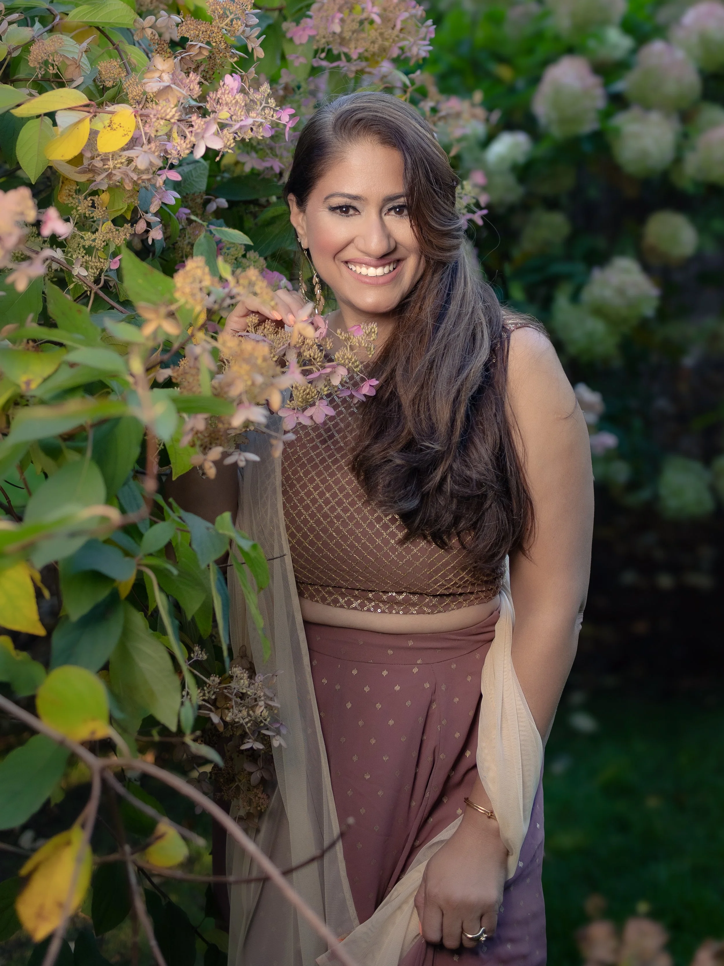 A woman with long, wavy brown hair and earrings smiling, standing amidst green foliage and pink hydrangea flowers, wearing a patterned top and a sheer shawl.