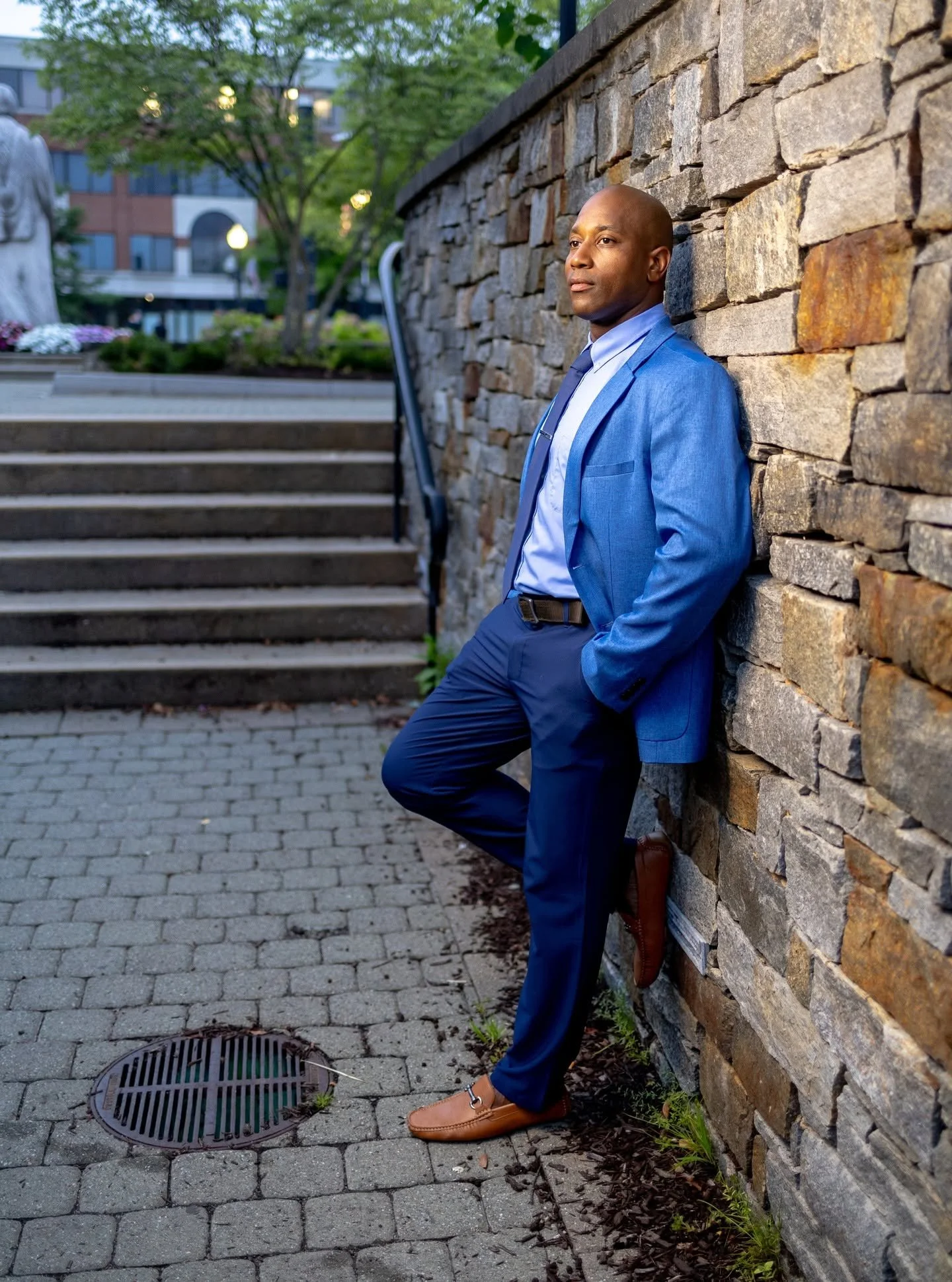 A man dressed in a blue suit with a white shirt and a dark tie, leaning against a stone wall with his eyes closed, outdoors in an urban setting with trees, flowers, and stairs visible in the background.