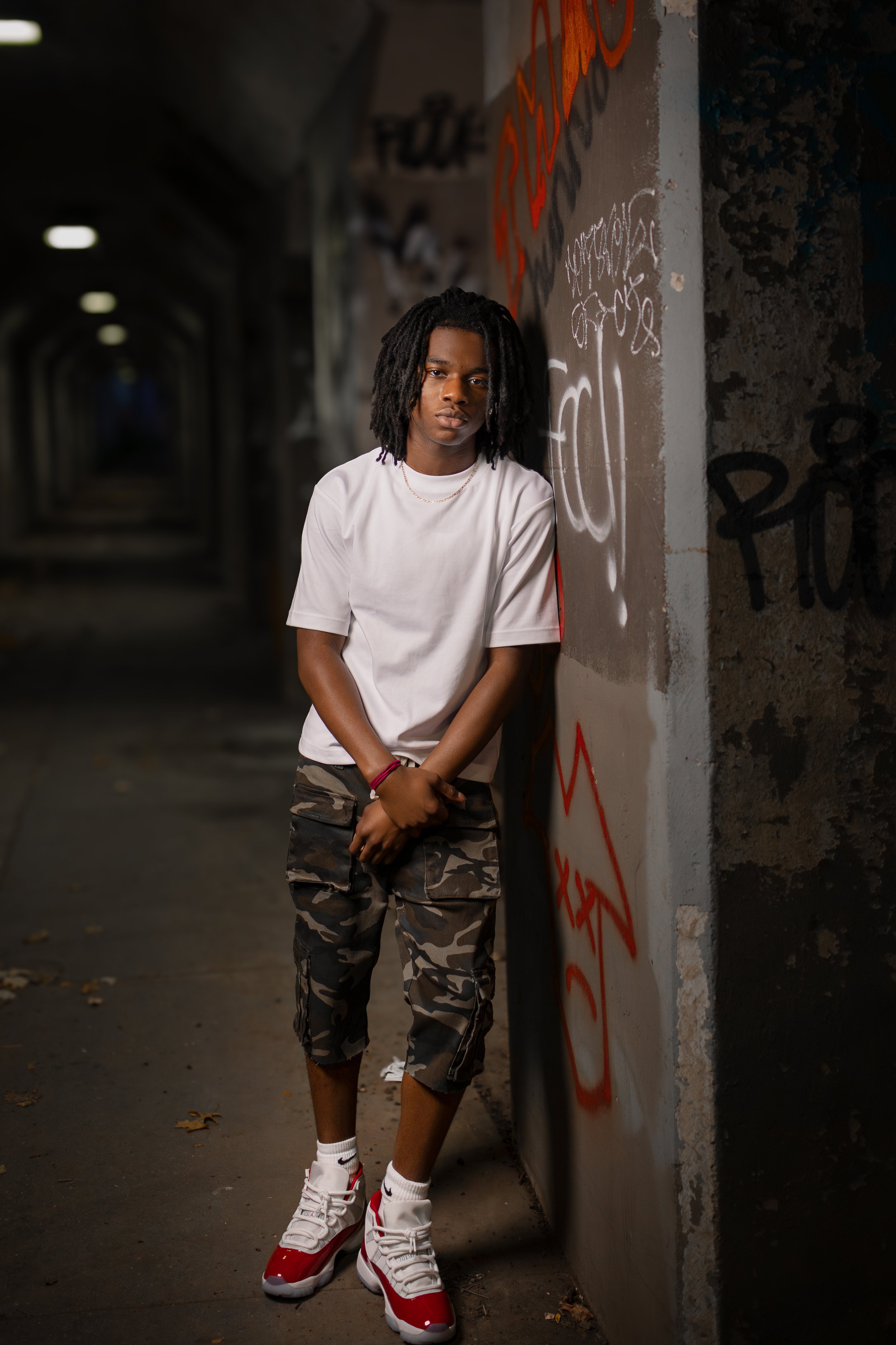 A young man with dreadlocks wearing a white t-shirt, camouflage shorts, and white sneakers with red accents standing in a graffiti-covered urban tunnel